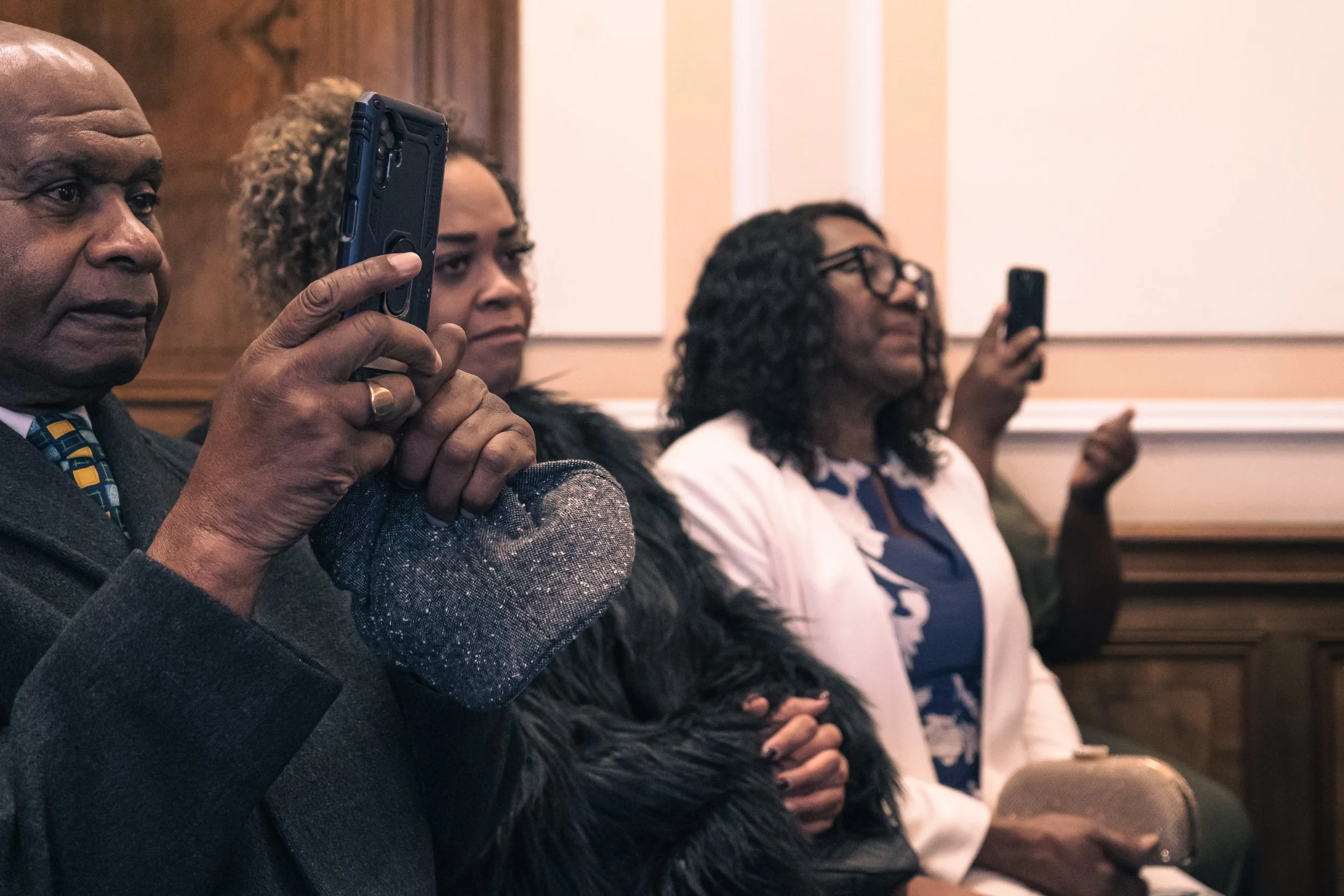 Three people sitting in a room, holding phones, appearing to take photos or videos. The man on the left is holding a phone with both hands, and the woman on the right is also holding a phone up. They are dressed in formal attire, and the setting appe