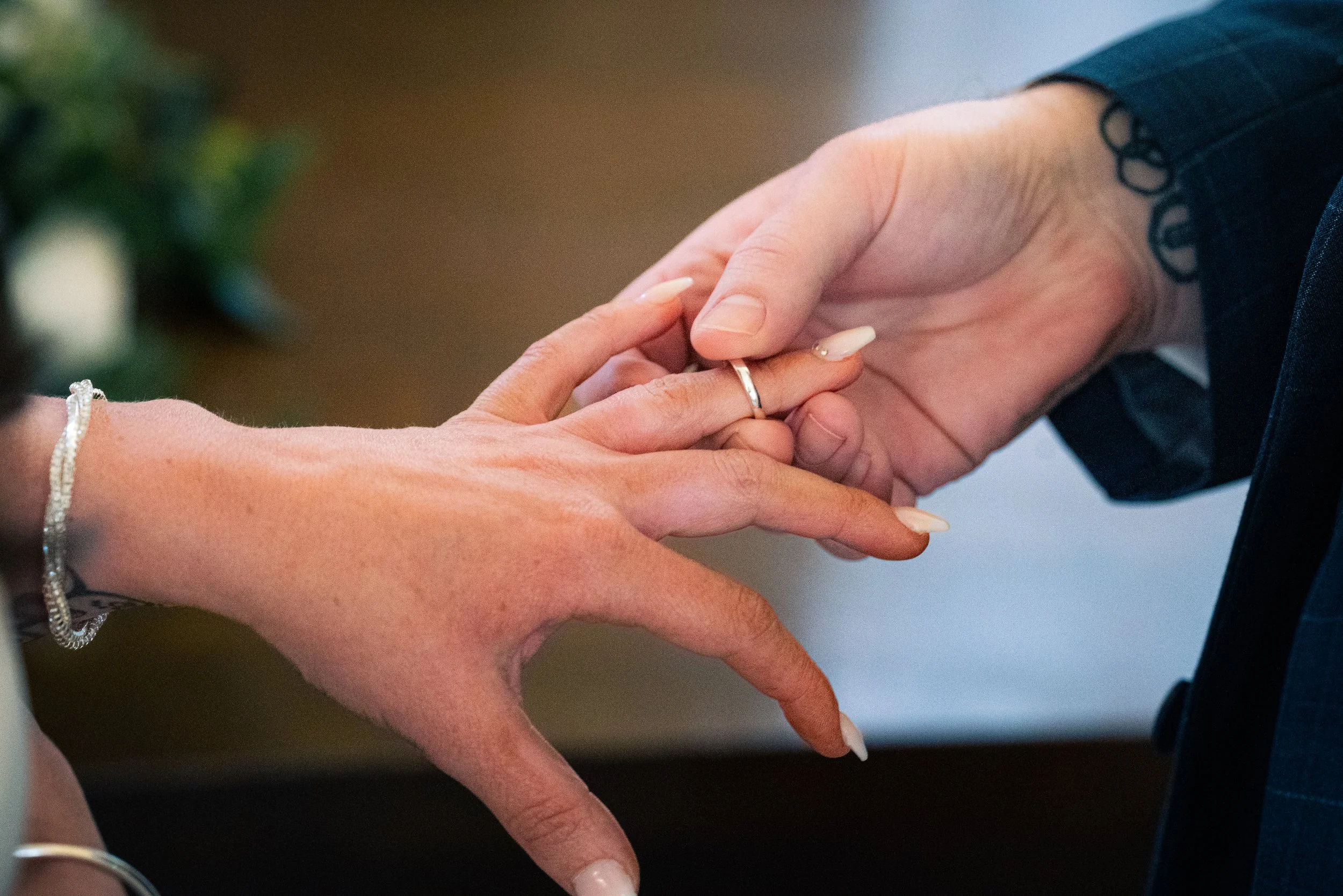 A close-up of a couple holding hands during a wedding ceremony, with the bride's hand over the groom's hand, showing a wedding ring, and both wearing jewelry.
