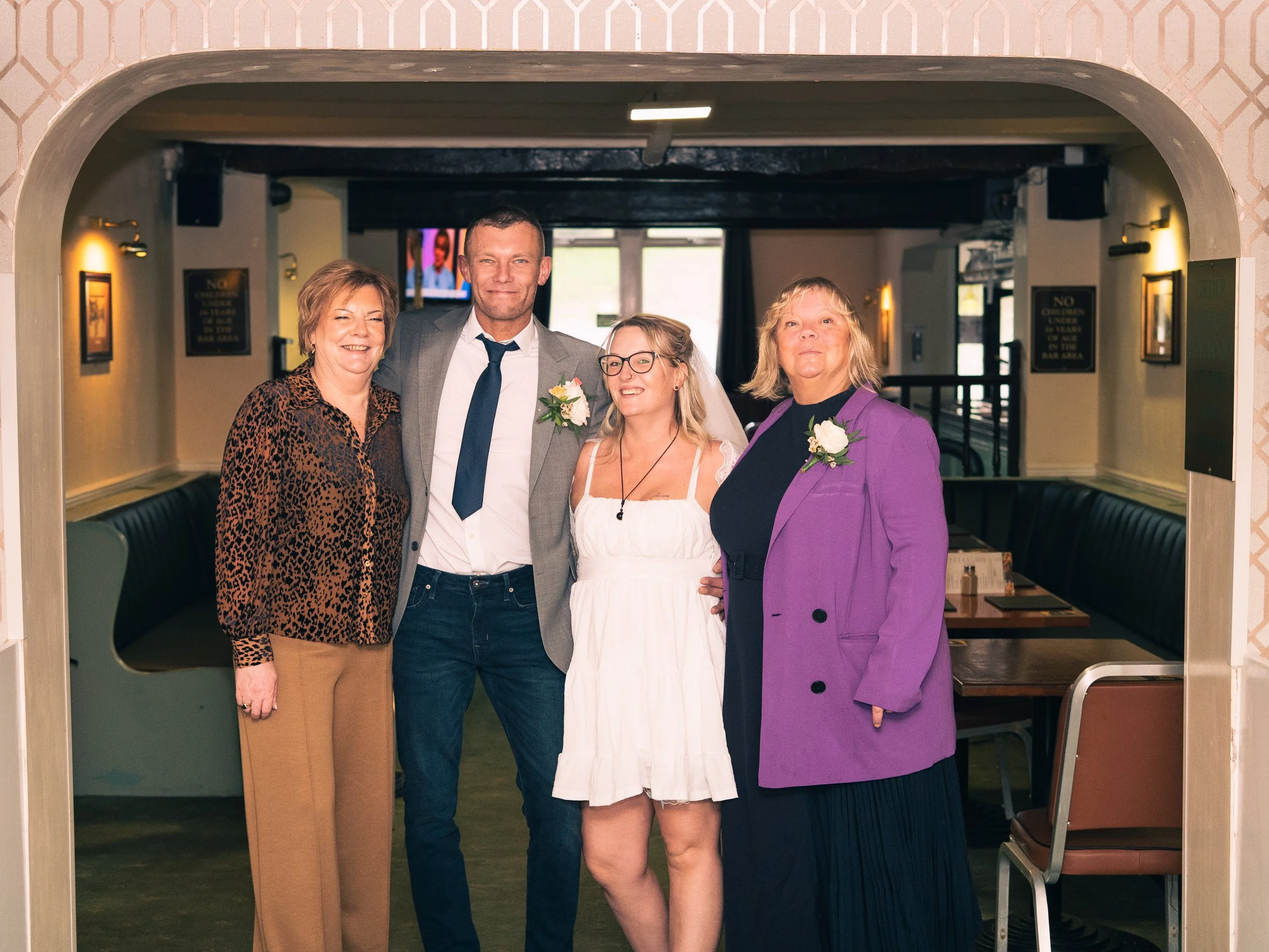 Four people dressed for a wedding, standing together inside a restaurant or banquet hall, smiling at the camera. The woman on the left is wearing a leopard print blouse and tan pants, the man next to her is in a gray suit with a white shirt and dark 