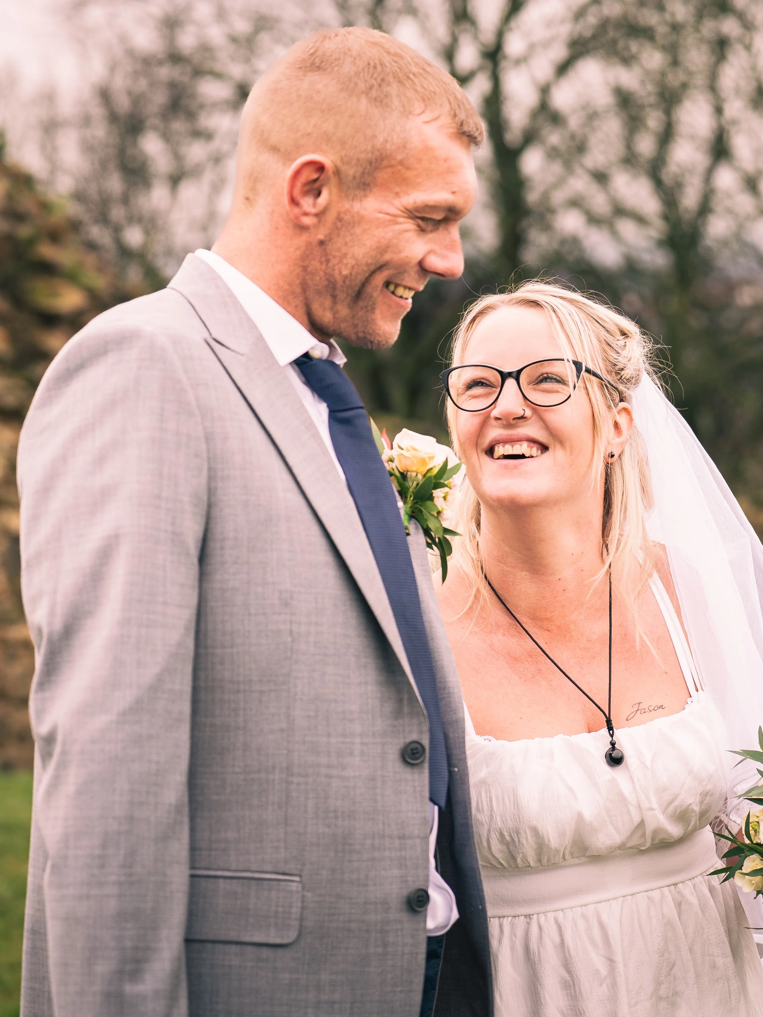 A smiling bride and groom on their wedding day outdoors, with the bride wearing glasses, a white dress, and veil, and the groom in a gray suit and blue tie.