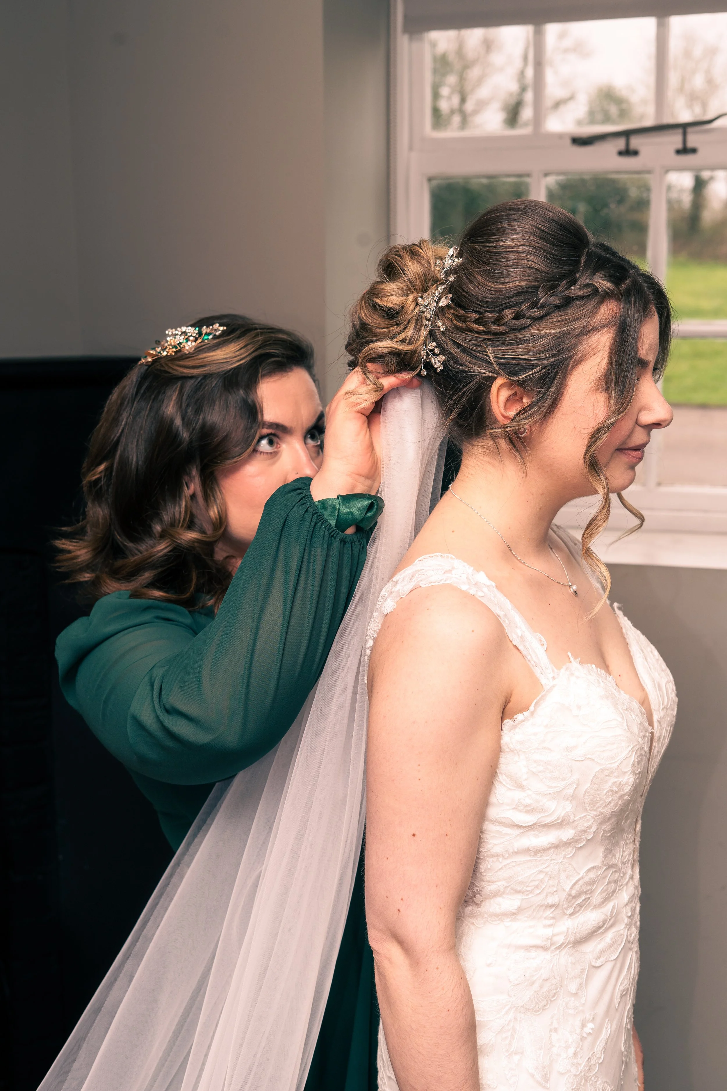 A bride in a white lace wedding dress with a veil is having her hair styled with the help of another woman, who is adjusting her veil.