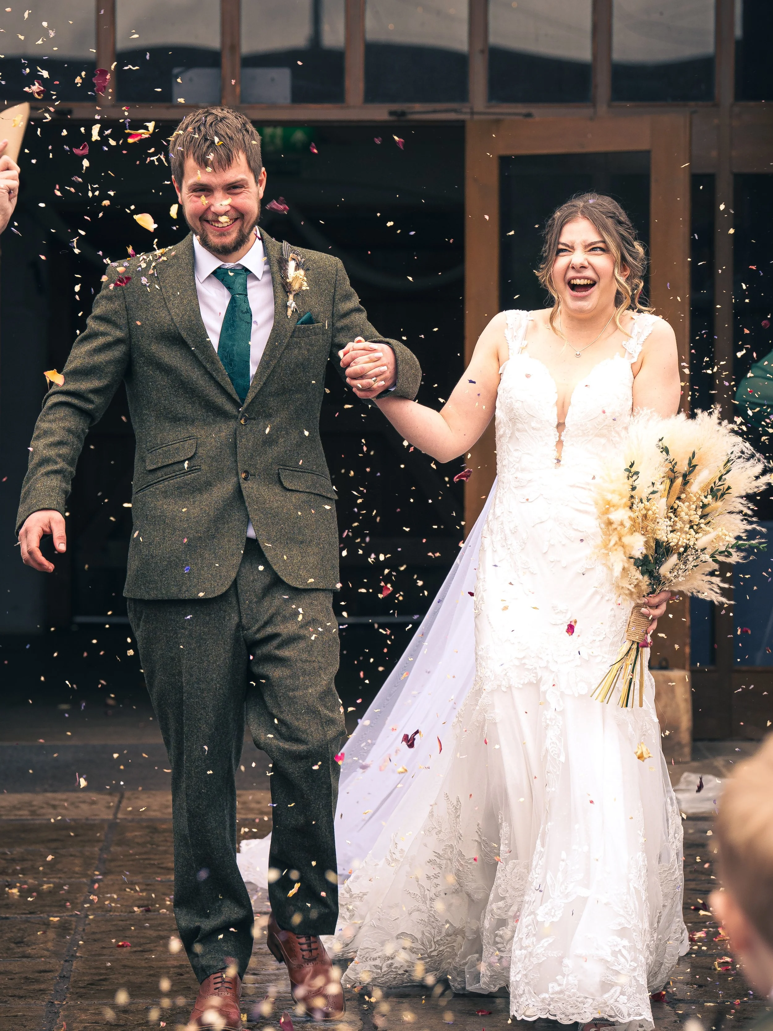 A newlywed couple holding hands and smiling as they walk through a shower of confetti during their wedding celebration outside a building. The bride is wearing a white wedding gown and holding a bouquet, while the groom is dressed in a suit.