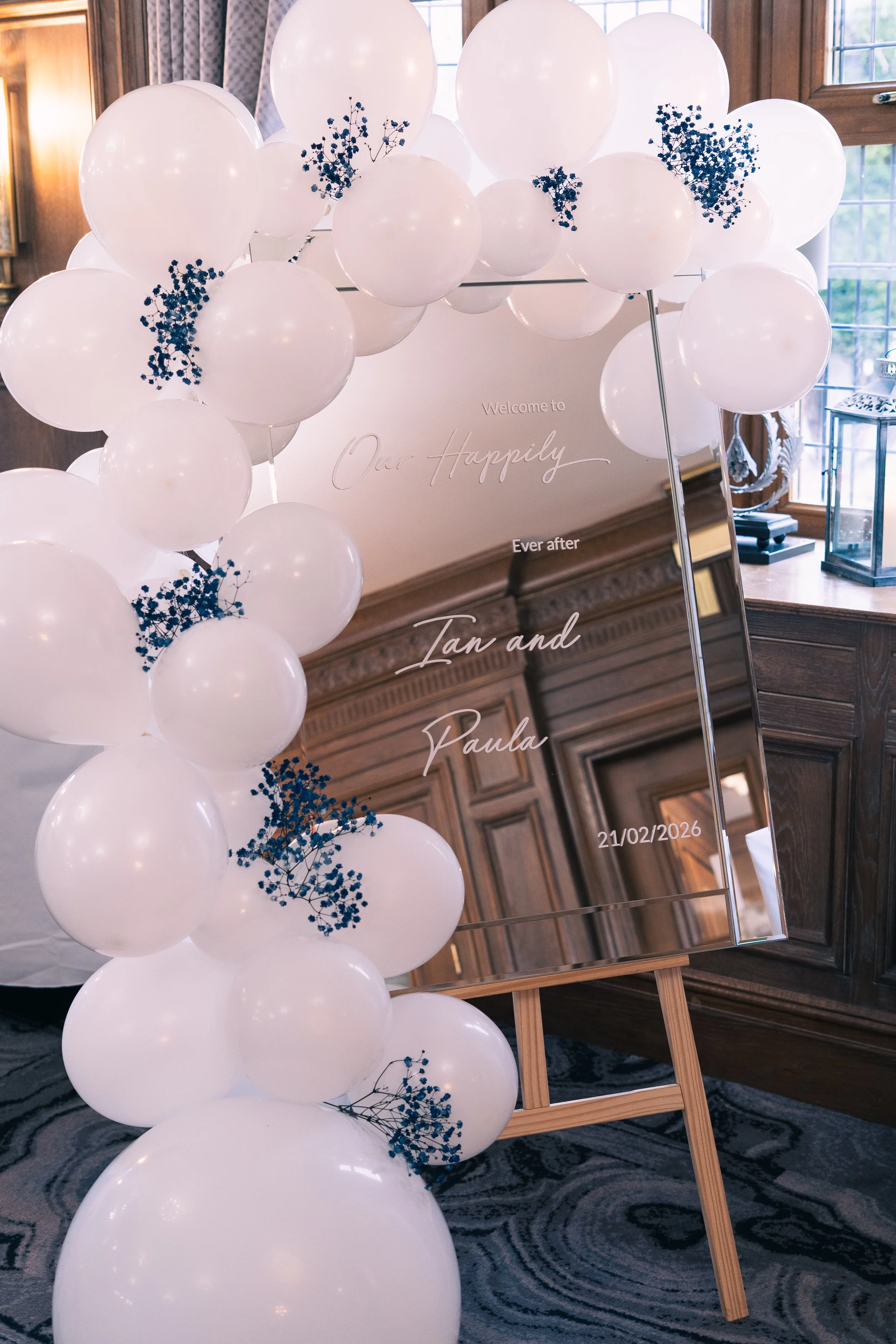 Decorative wedding sign framed by white balloons with small blue flowers, displaying the names Tan and Paula and the date 21/02/2026.