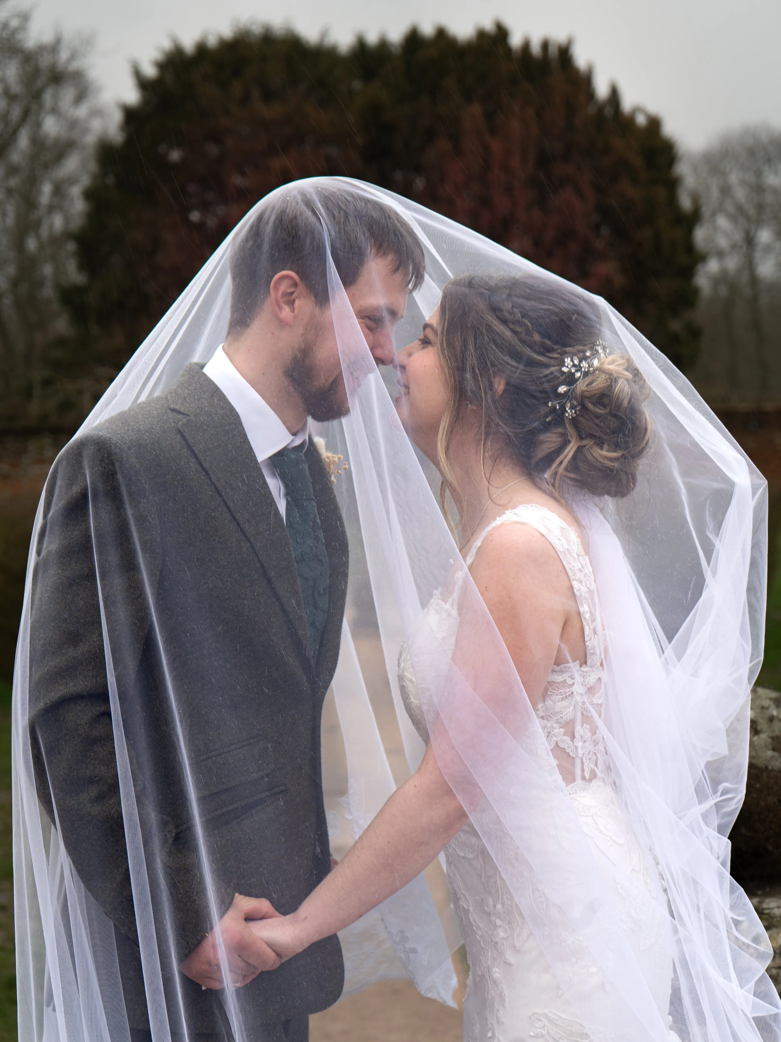 A bride and groom holding hands and leaning in close with a veil covering them, outside in a natural setting with trees.