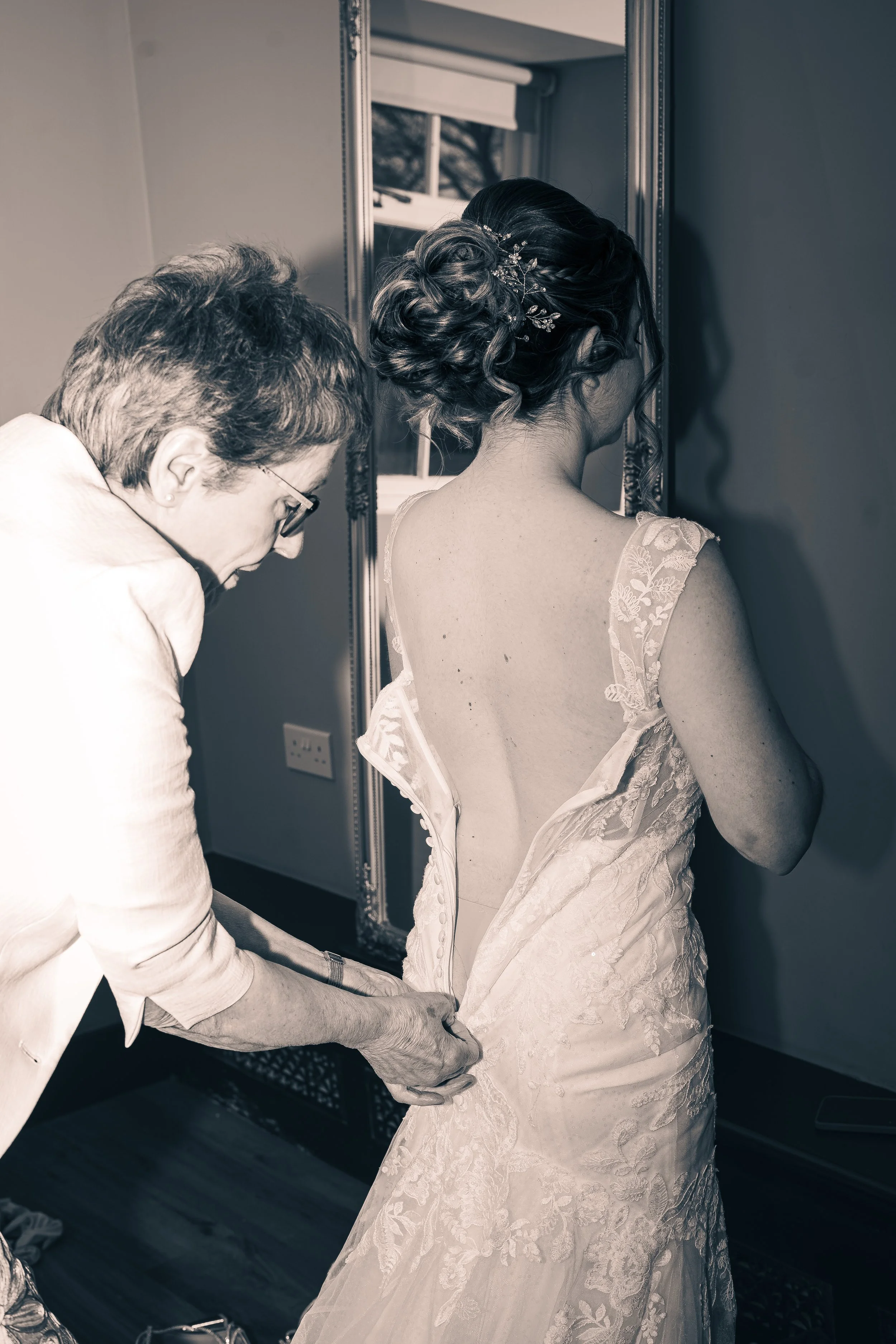 A woman in a wedding dress having her gown adjusted by another woman, with a mirror and window in the background.