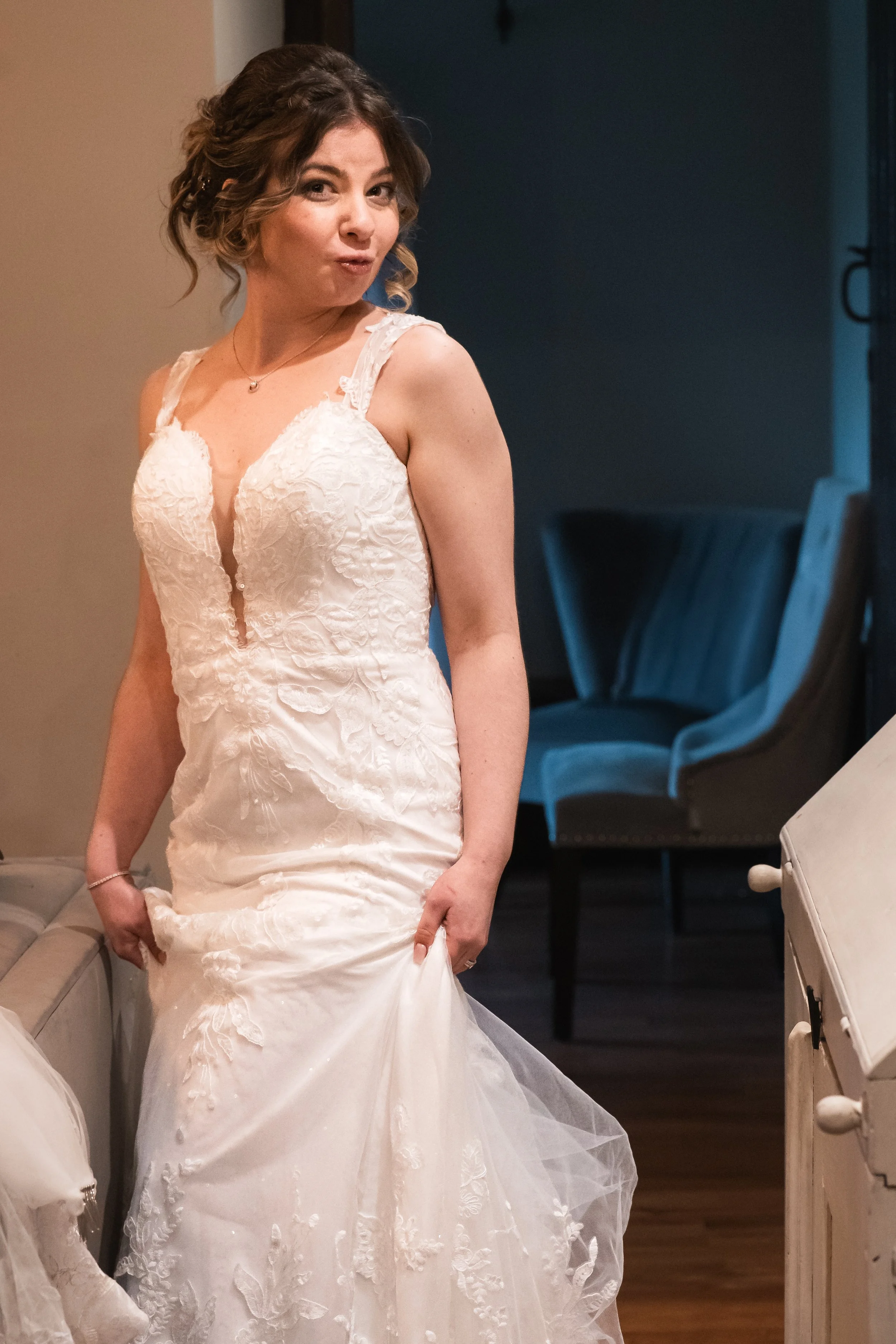 A woman in a wedding dress standing indoors, holding the hem of her dress, with a surprised or playful expression.