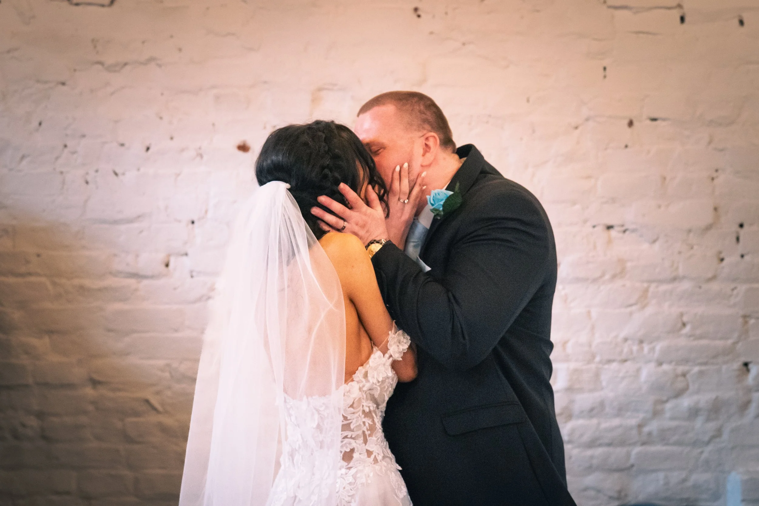 A couple sharing a kiss during their wedding, with the bride wearing a lace wedding gown and veil, and the groom in a black tuxedo with a blue boutonniere, against a white brick wall.