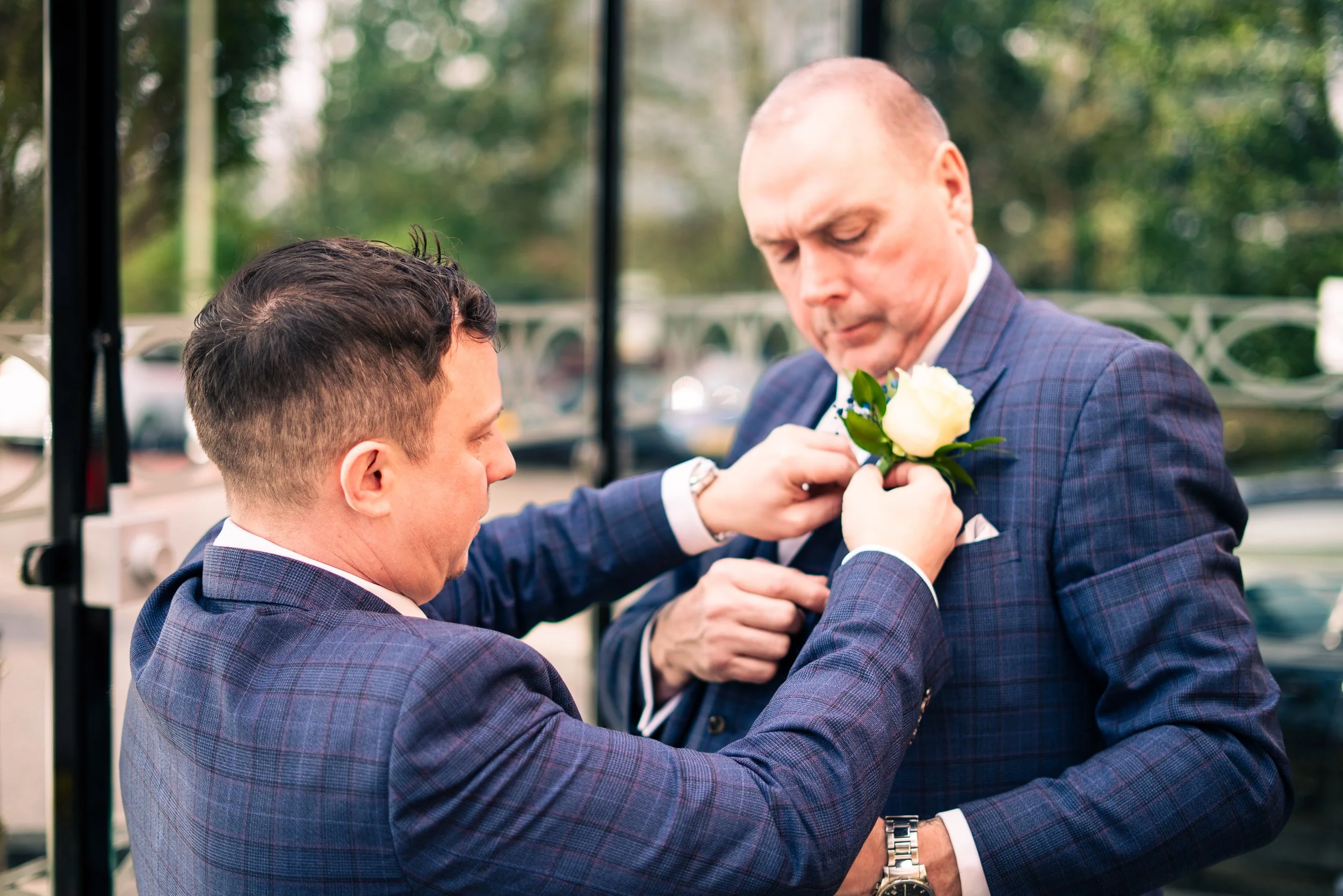 Two men in suits, one younger and one older, dressed in checked suits, with the younger pinning a white rose boutonniere onto the older man's lapel outdoors.