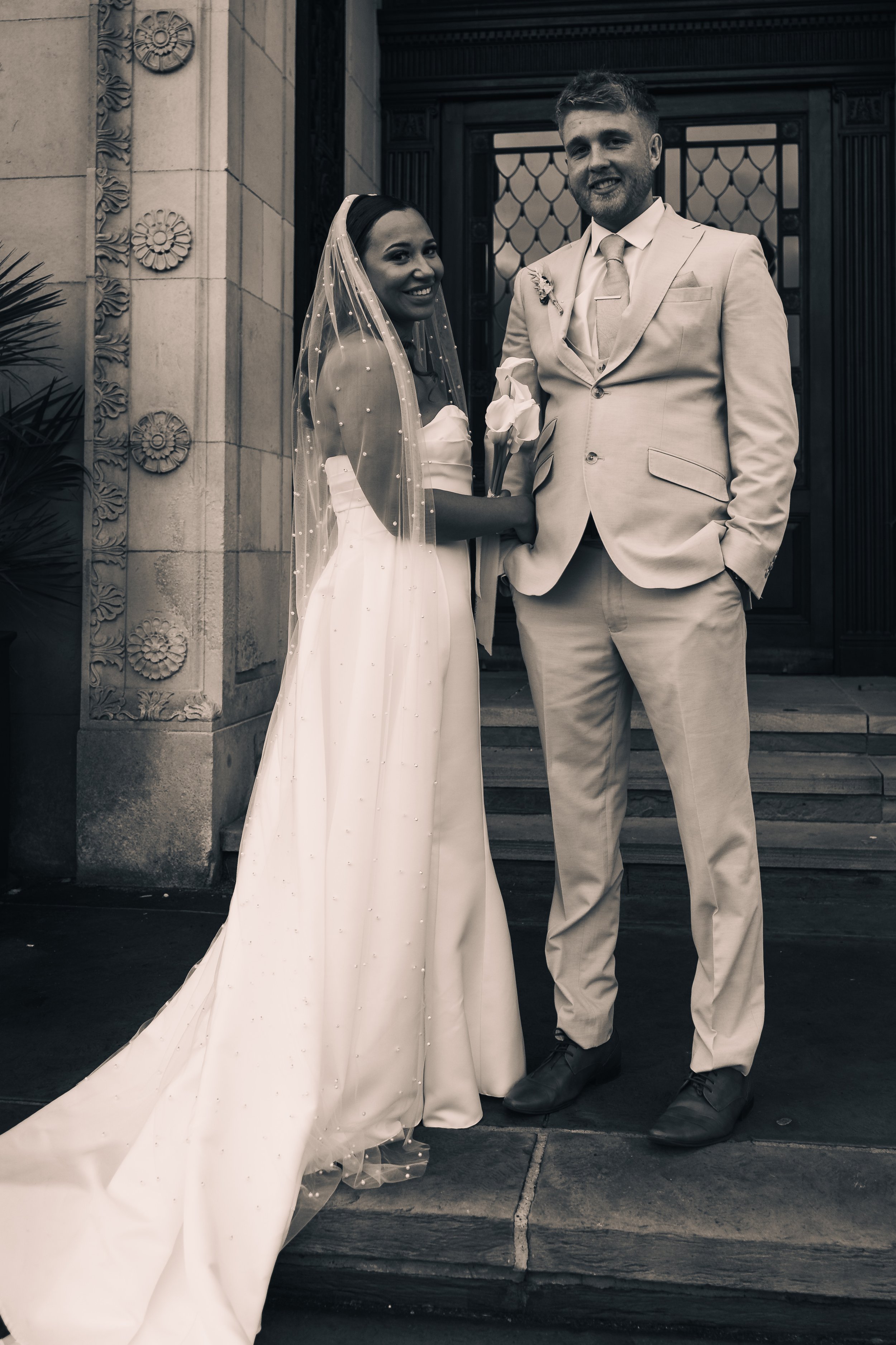 A bride and groom standing outside a building, smiling for a wedding photo. The bride wears a white wedding dress with a veil, holding a white calla lily. The groom wears a light-colored suit with a tie, with one hand in his pocket.