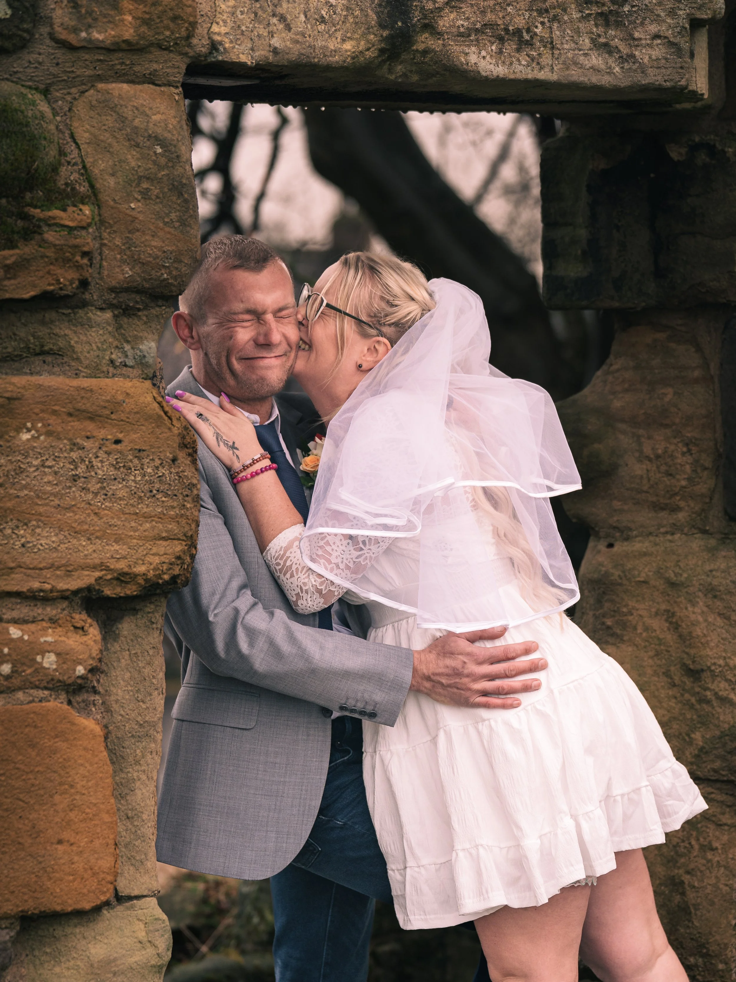 A couple sharing a kiss through a stone arch, the woman wearing a white dress and veil, the man in a gray suit with a boutonniere, outdoors with trees in the background.
