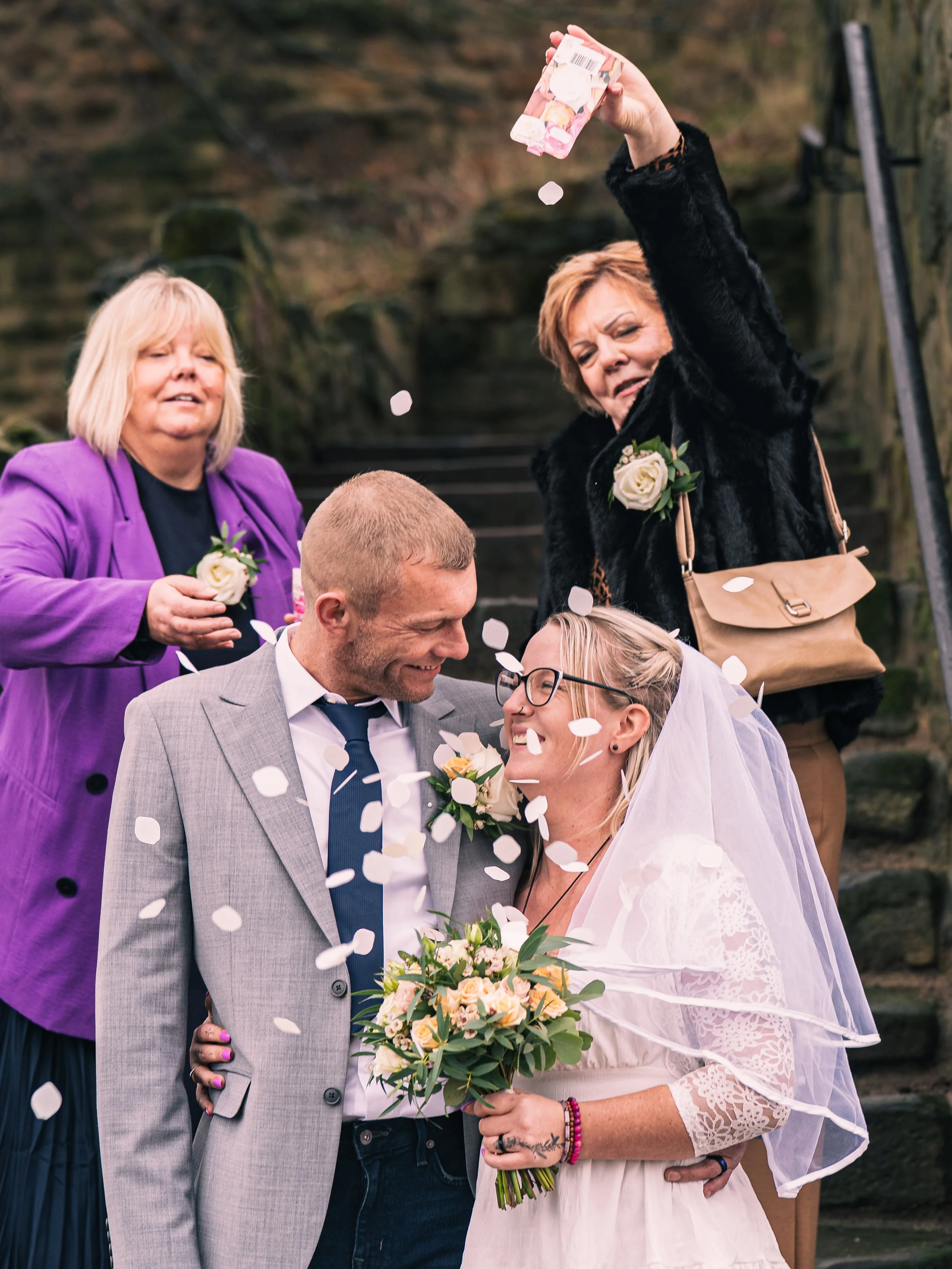 A bride and groom celebrate their wedding with confetti being poured over them, surrounded by friends and family on outdoor stone steps.