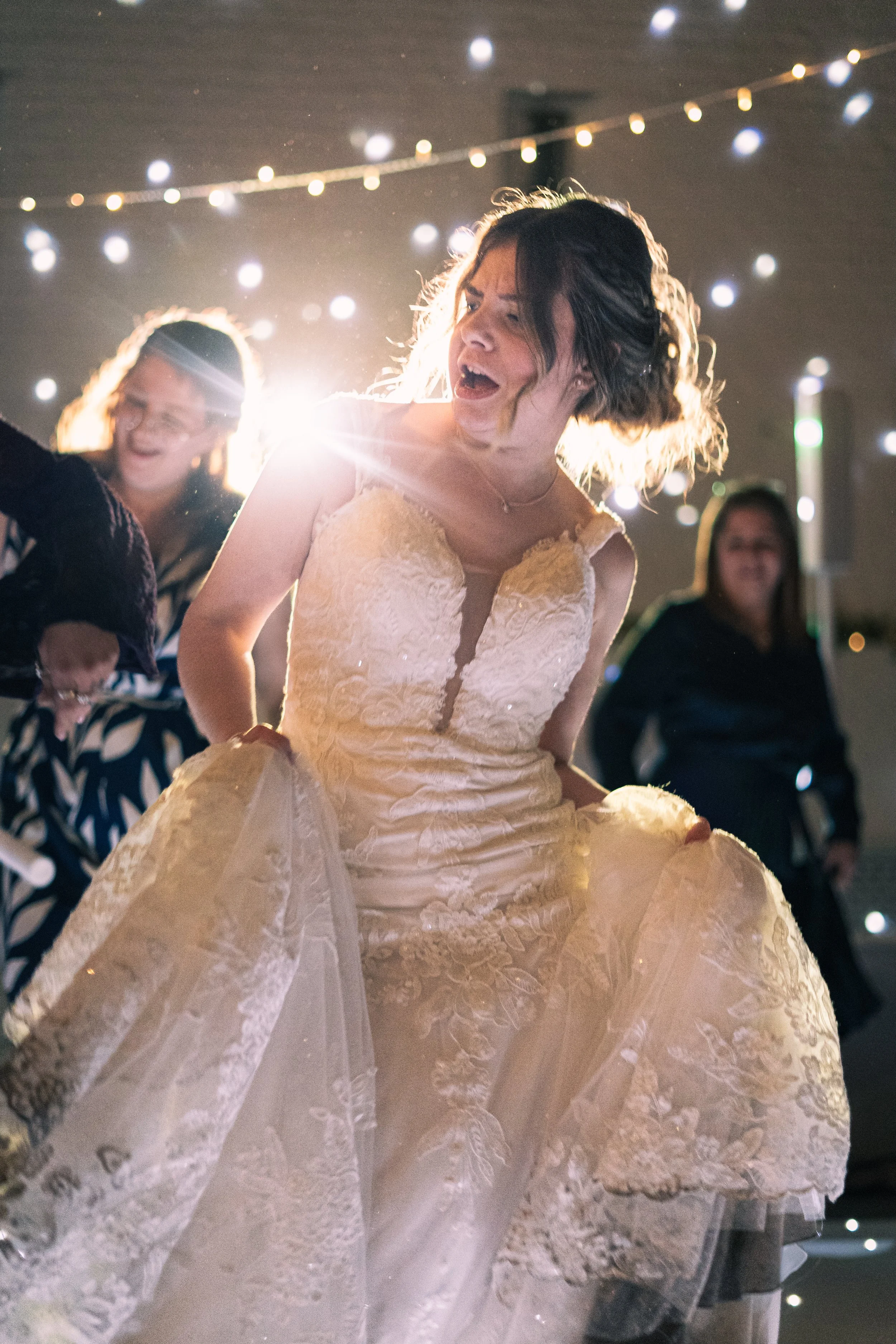 Woman in a wedding dress dancing joyfully at a celebration, with friends in the background and string lights overhead.