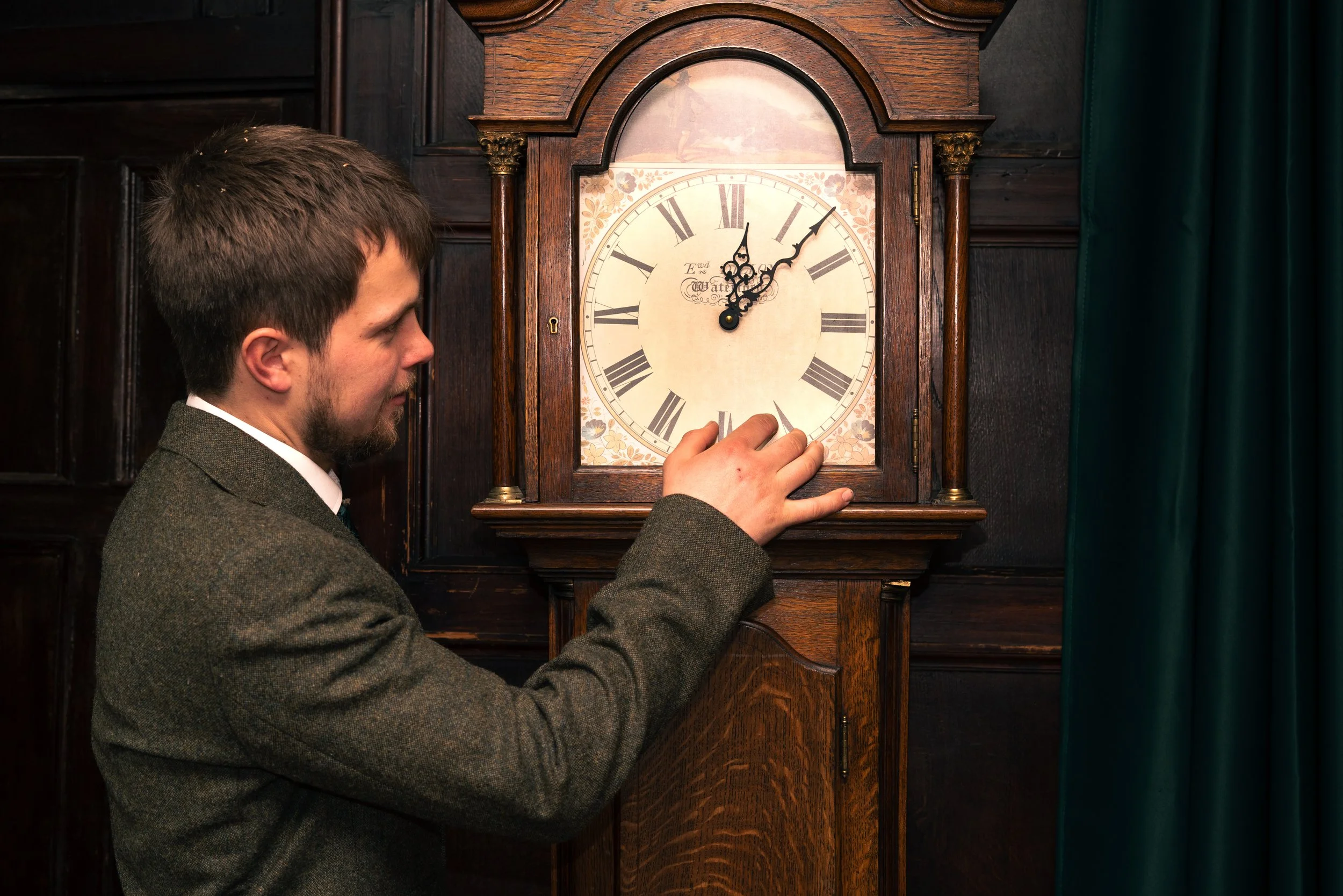 A man in a brown suit adjusting the pendulum of a vintage wooden grandfather clock with a white face, Roman numerals, and ornate black hands inside a wood-paneled room.