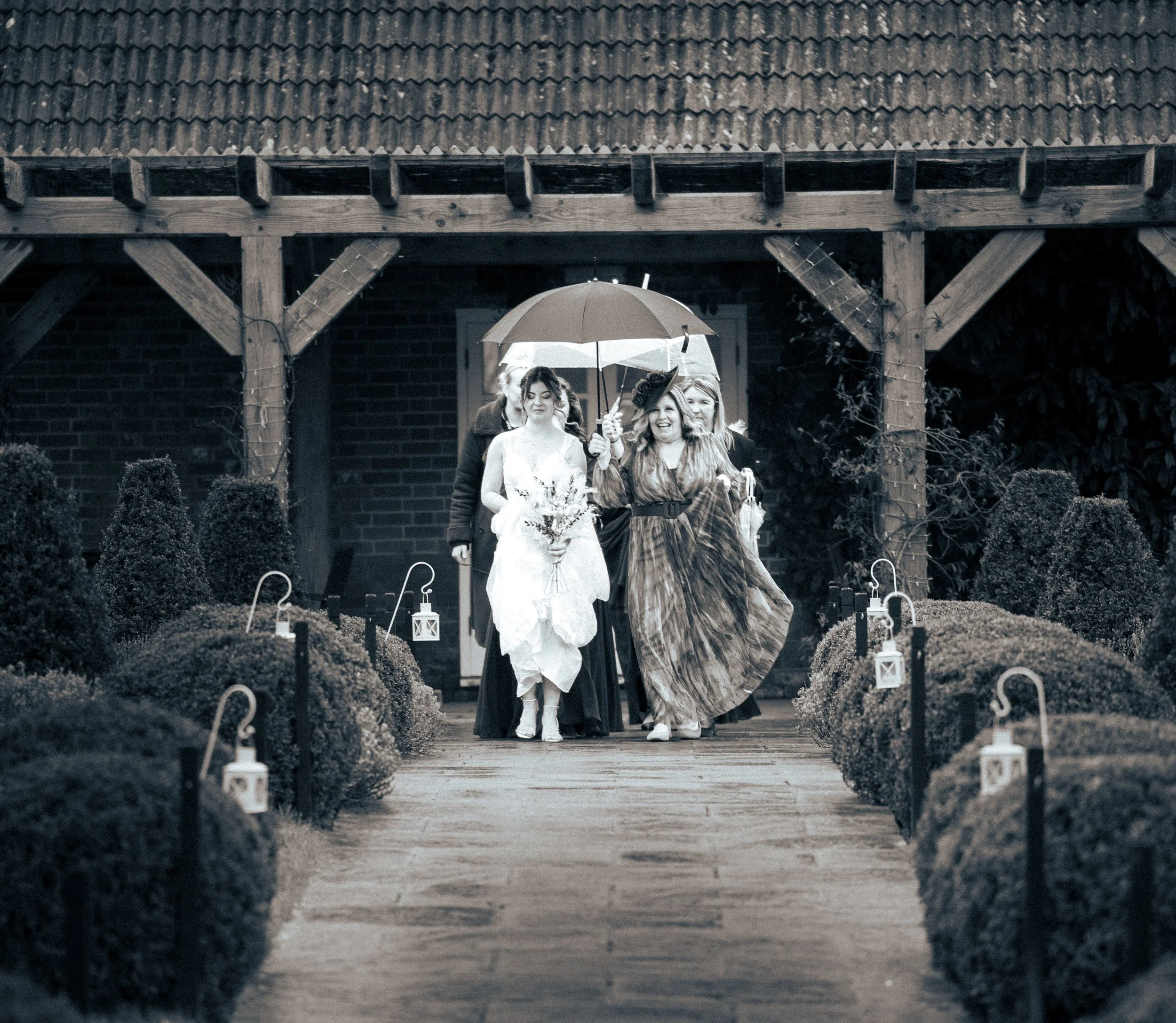 People walking down a garden path holding an umbrella, with shrubbery and lanterns along the sides.