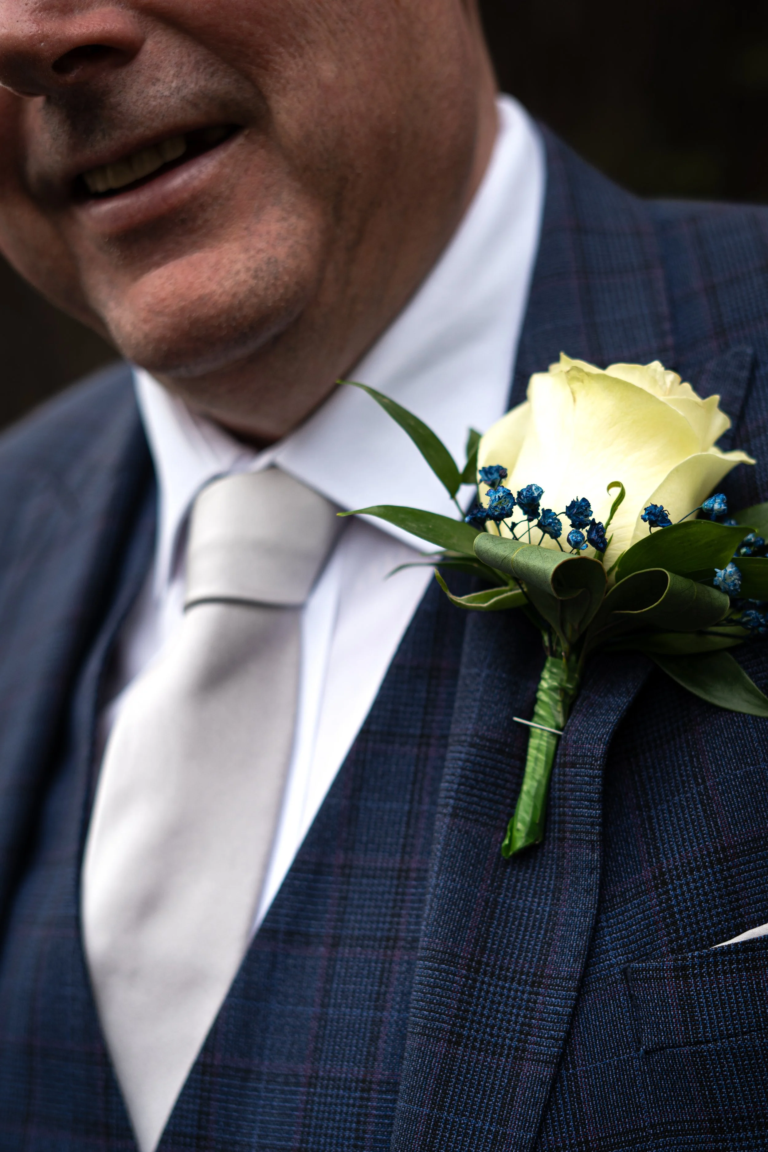 Close-up of a man in formal attire with a white tie and dark plaid suit, wearing a boutonniere with a white rose and small blue flowers on his lapel.