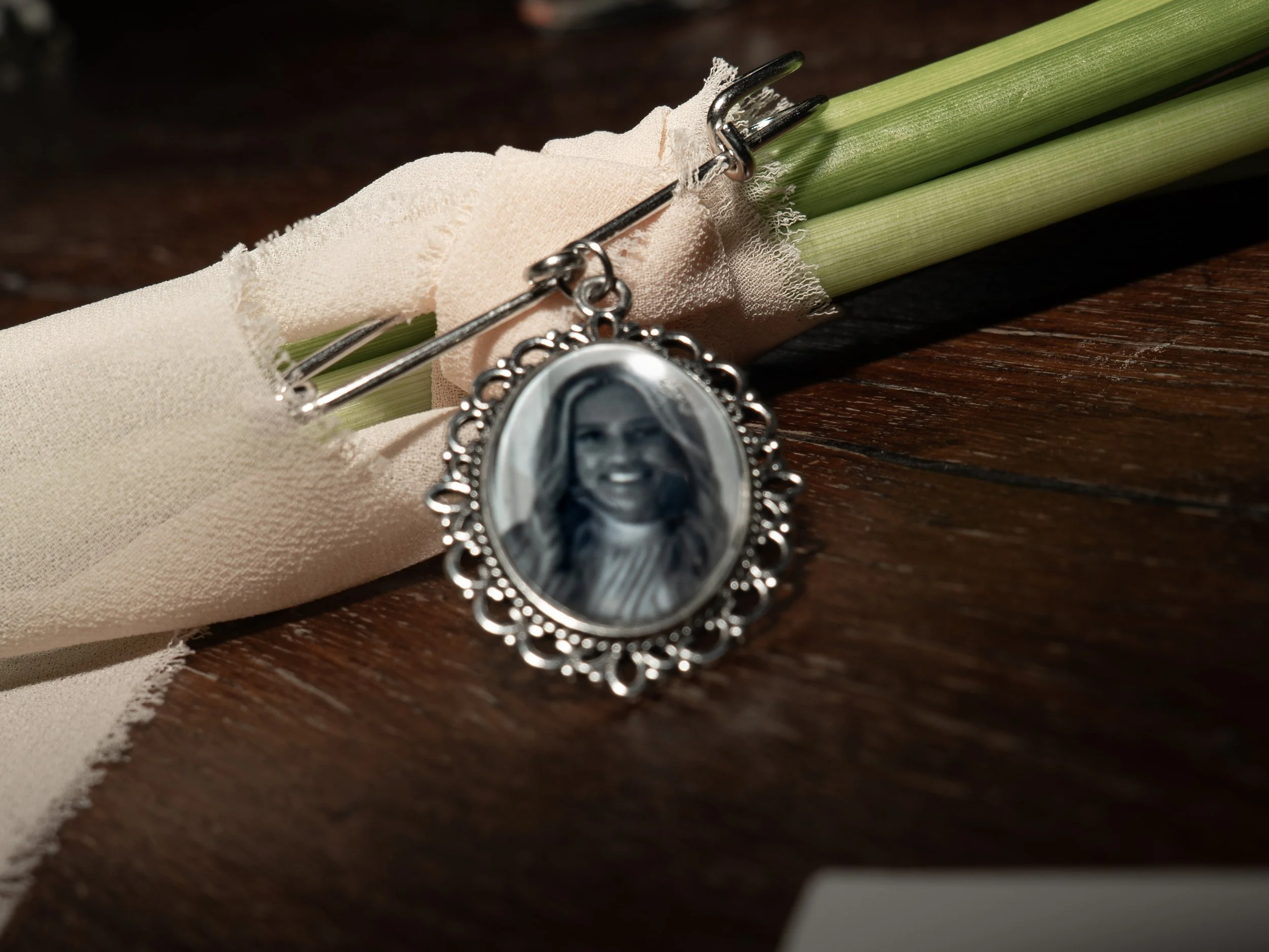 A rosary with a silver pendant depicting a woman's face and a bunch of green flowers wrapped in beige cloth, placed on a wooden surface.