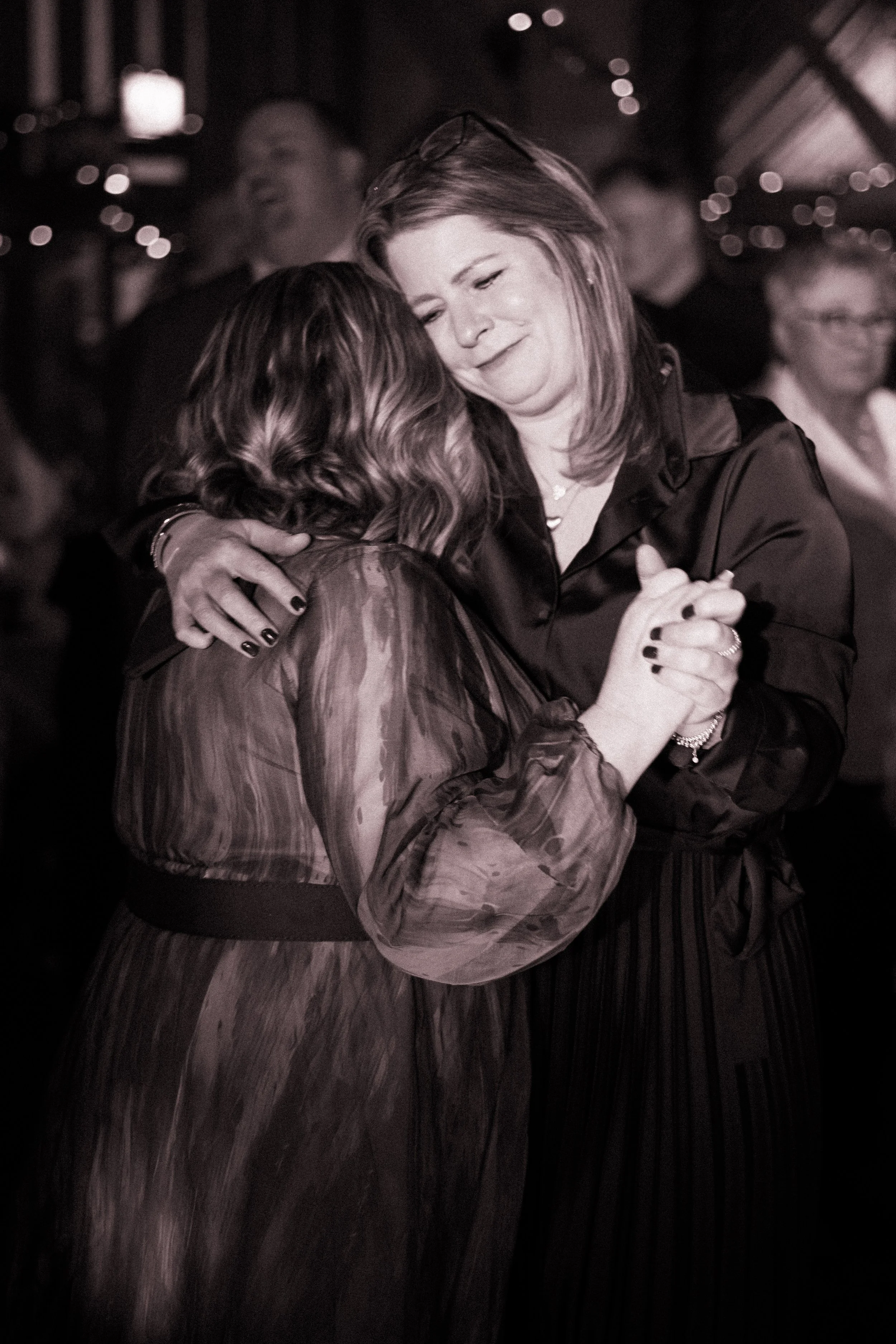Two women are dancing closely, embracing each other, in a dimly lit setting. The woman facing the camera has light-colored hair and is smiling gently with her eyes closed, while the other woman has dark, wavy hair and is facing away from the camera.