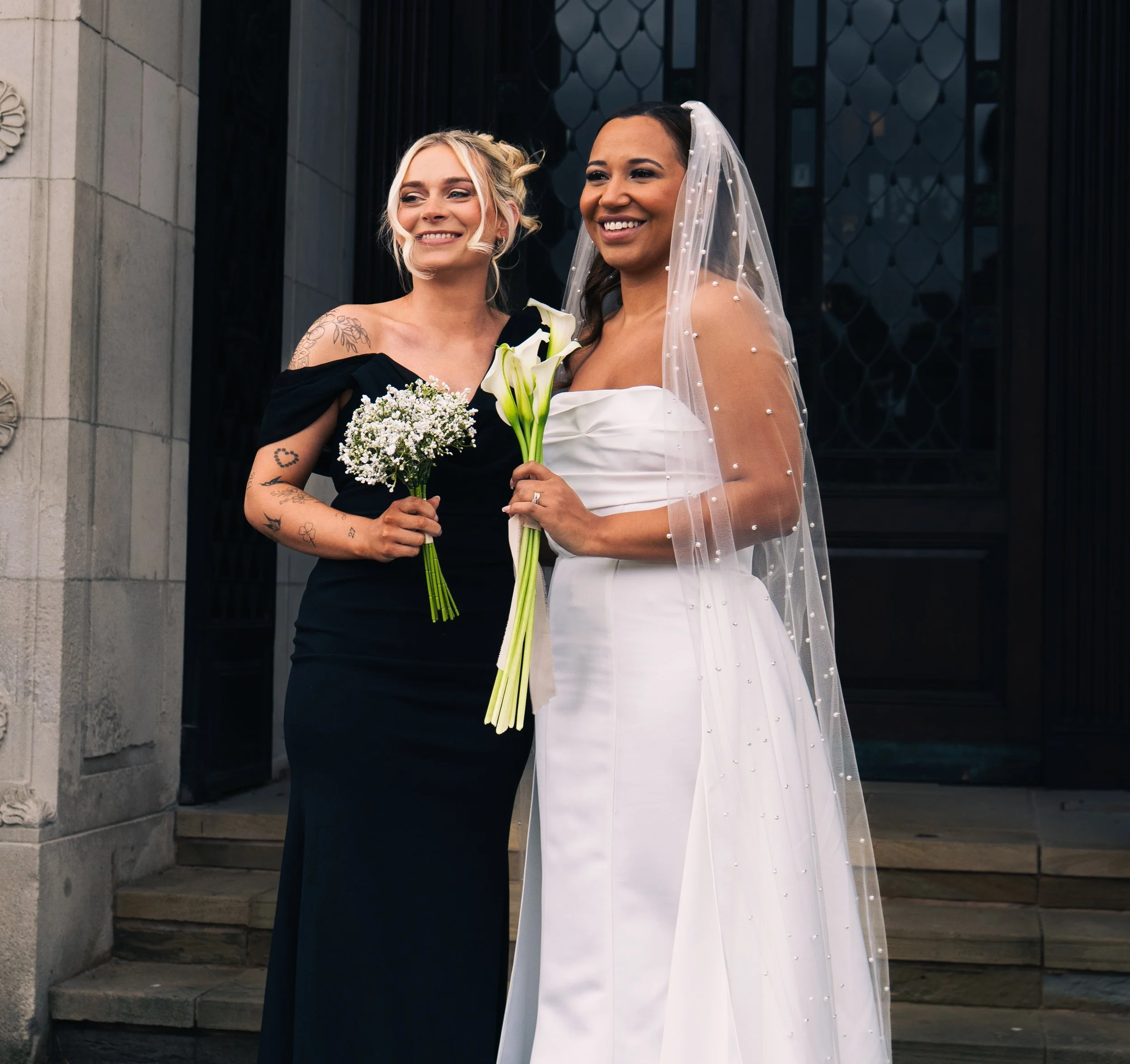 Two women standing in front of a building, one in a black dress holding a small bouquet, and the other in a white wedding dress with a veil holding a bouquet of white calla lilies and flowers, smiling.