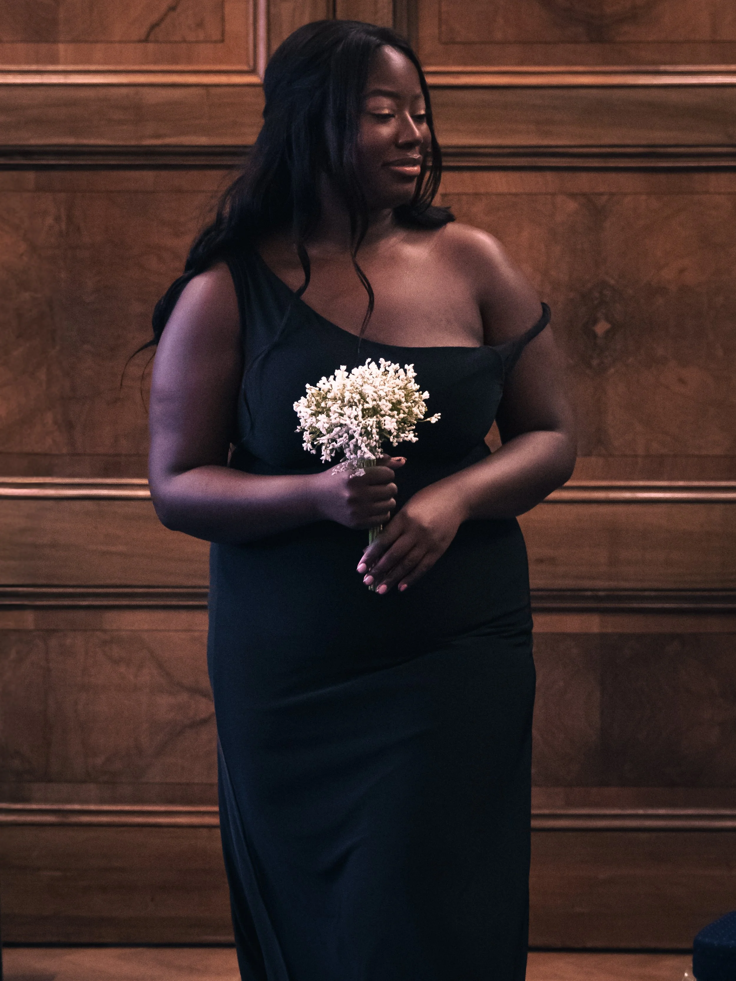A woman with long black hair is wearing a black dress with one shoulder strap and is holding a bouquet of small white flowers, standing against a wooden-paneled wall.