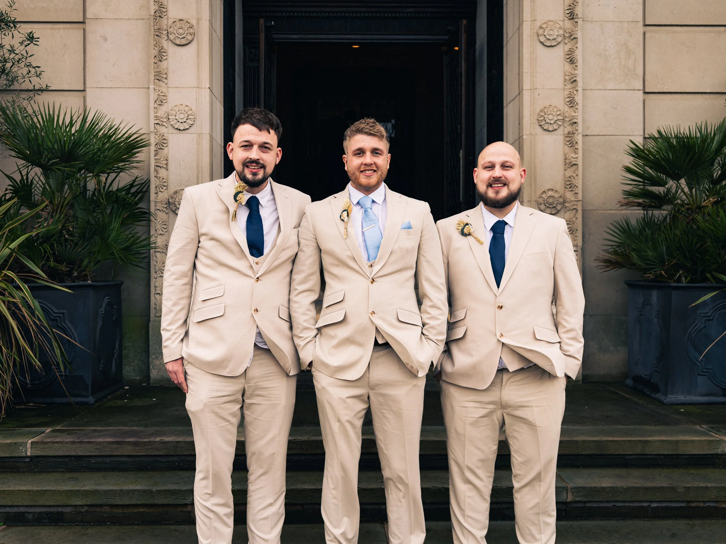 Three men dressed in light beige suits with dark blue ties and boutonnières, standing on stone steps in front of a building with decorative stonework and potted plants, smiling at the camera.