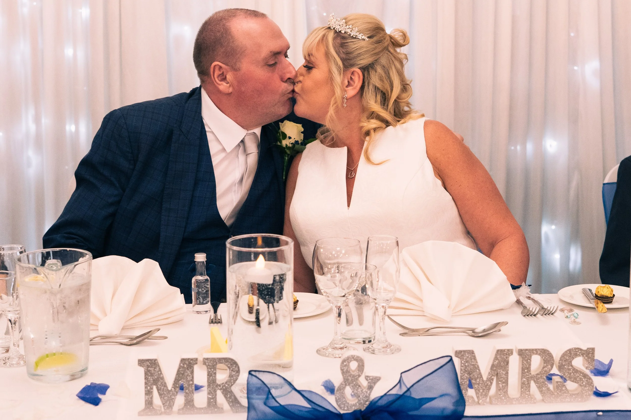 A bride and groom kiss at their wedding reception, sitting at a decorated table with candles and initials 'MR & MRS' in front of them.