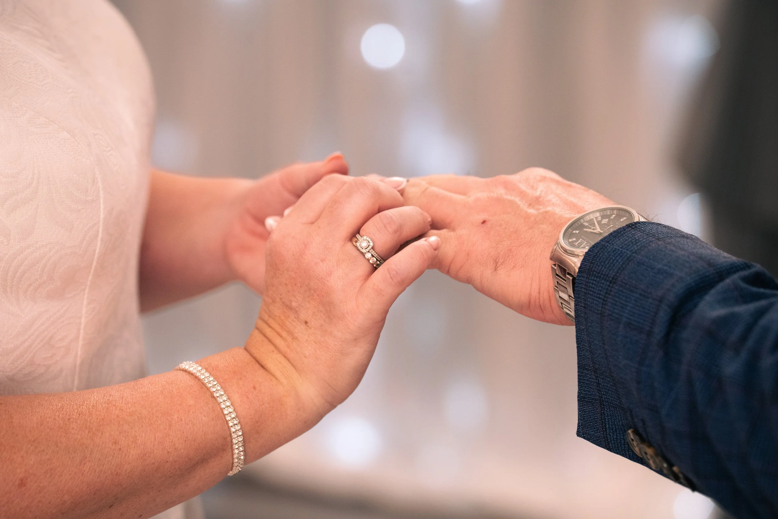 A woman and a man holding hands, with the woman's hand resting on top of the man's hand. The woman is wearing a diamond ring and a diamond bracelet, while the man is wearing a watch with a metal band. The background is blurred with some circular ligh