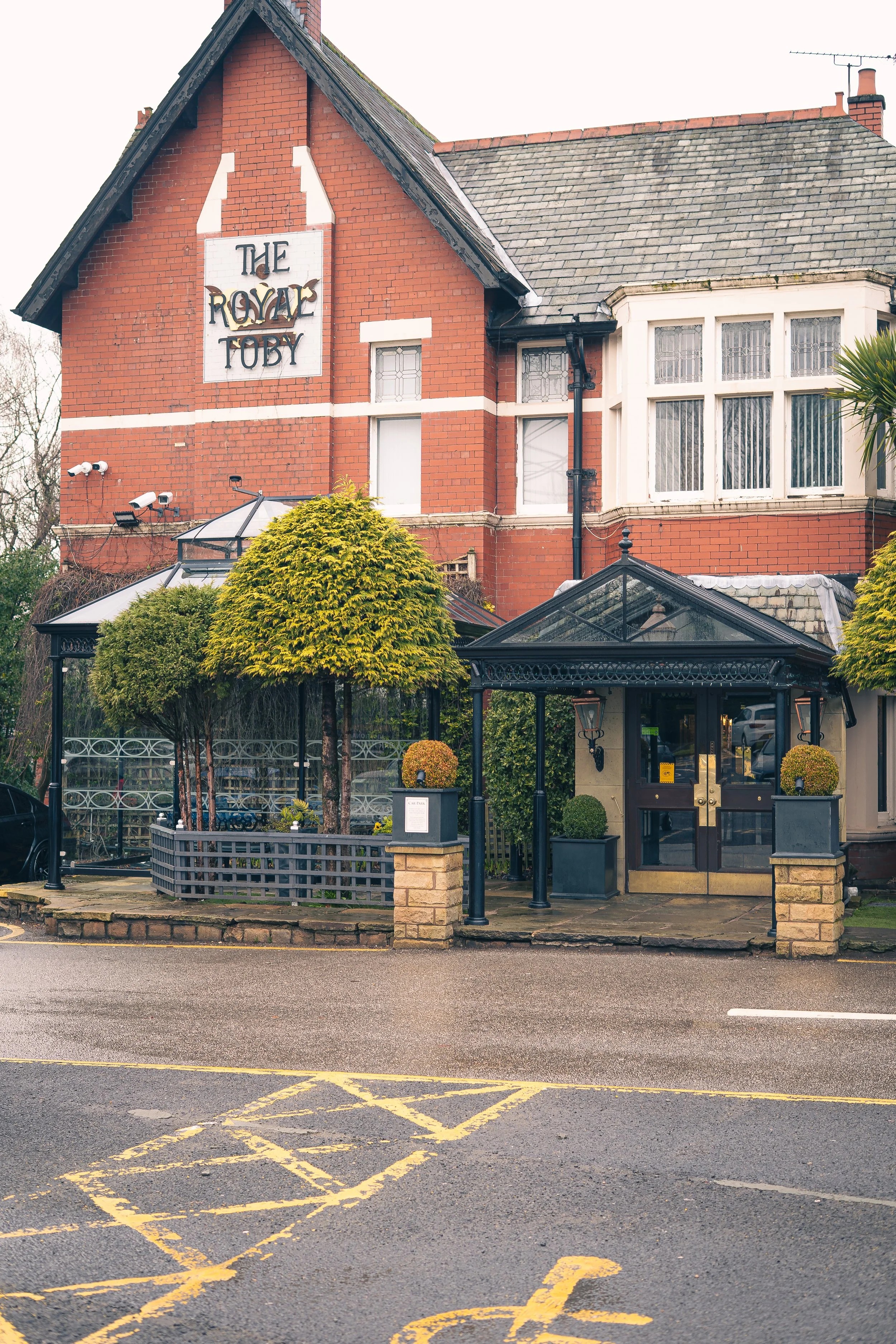 Exterior of a brick building with a sign that reads 'The Royal Toby.' The entrance has a glass door under a black metal covered porch, and landscaped with trimmed trees and potted plants.