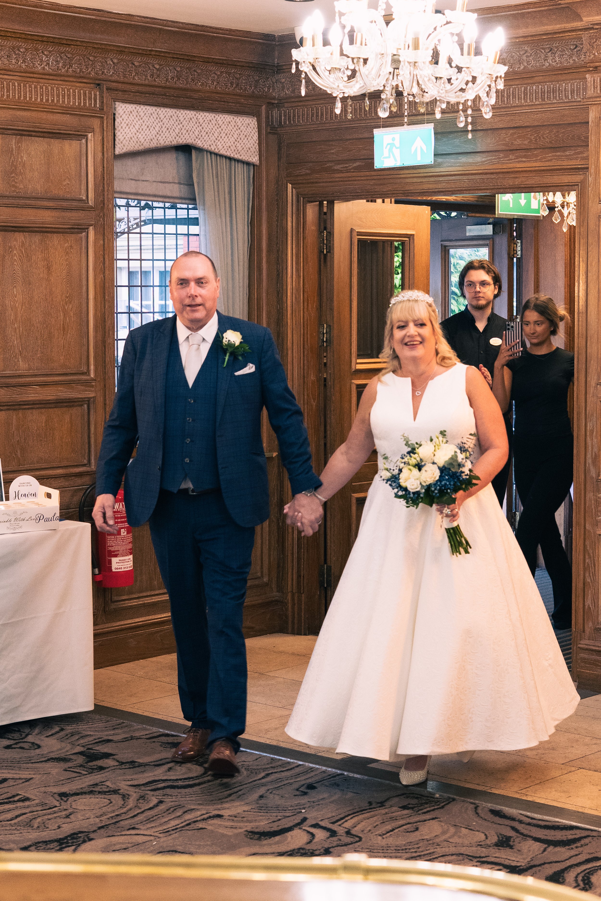 A bride and groom holding hands as they walk into a room for their wedding celebration. The bride is wearing a white wedding gown and holding a bouquet of flowers, while the groom is dressed in a blue suit with a white boutonniere. They are smiling a