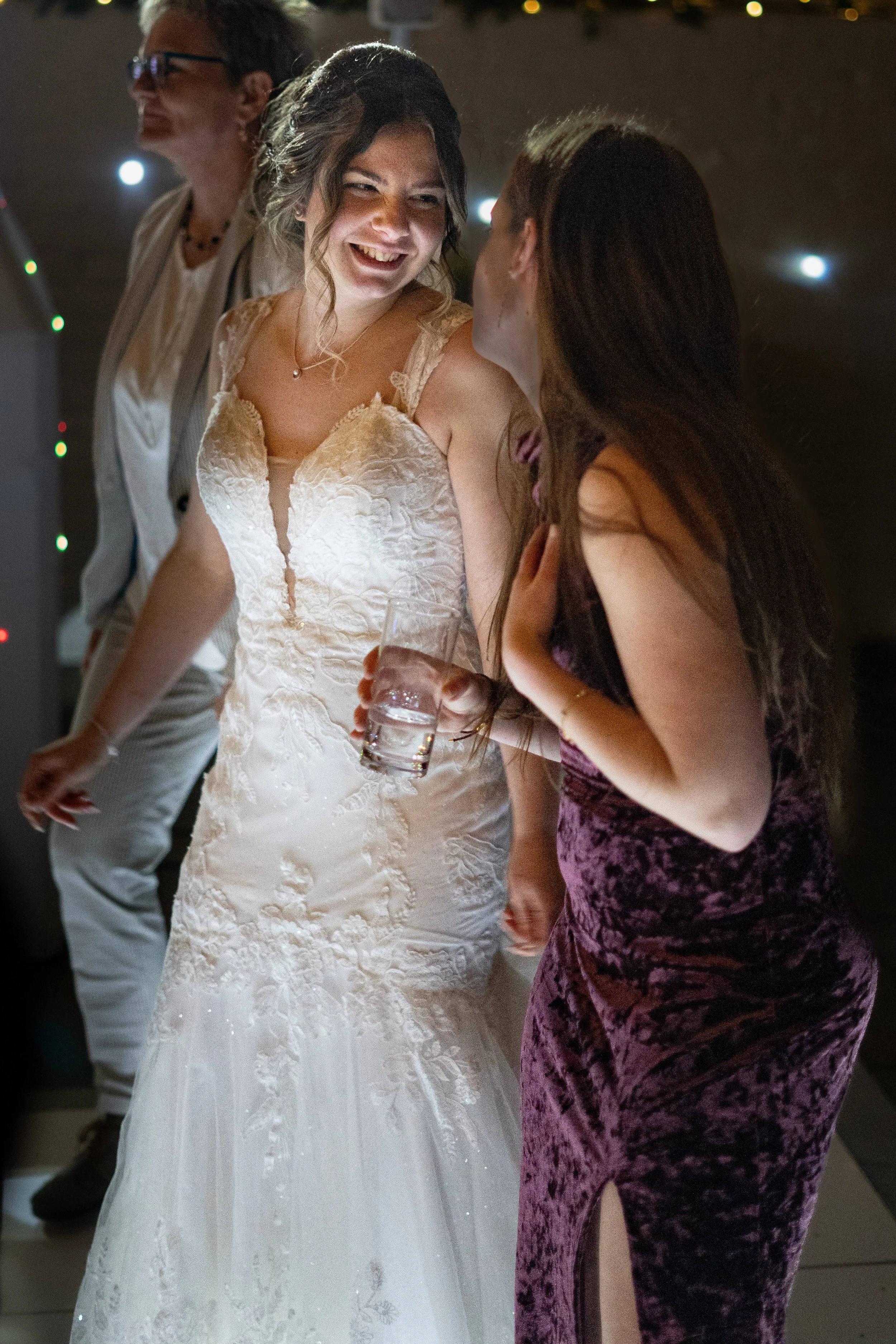A bride in a white wedding dress smiling and talking to a woman in a purple velvet dress, with two other people, a man and a woman, in the background. The bride is holding a glass of water.