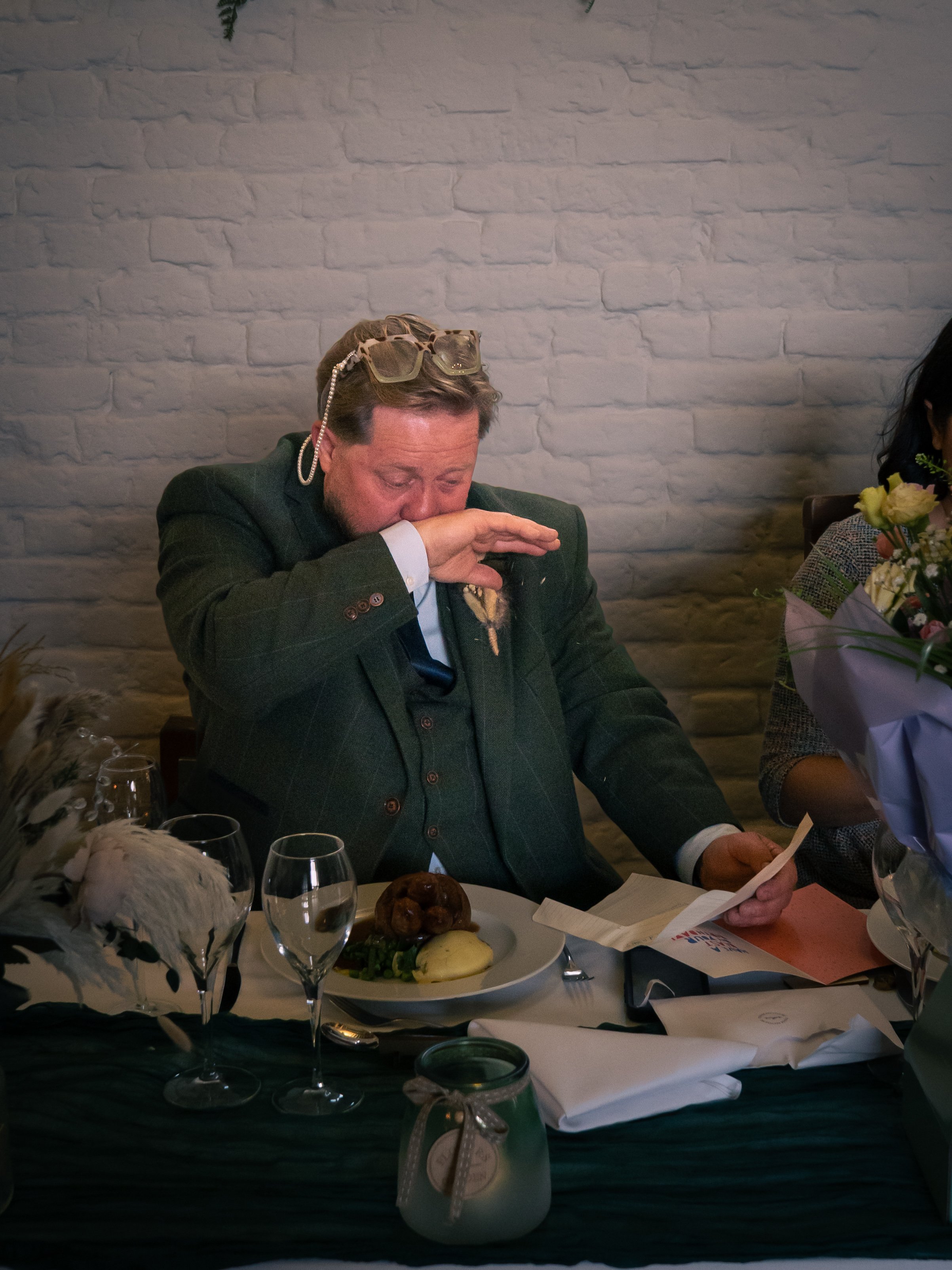 A man in a dark green suit with glasses on his head is wiping tears while holding a piece of paper, sitting at a table with food, glasses, flowers, and a candle.