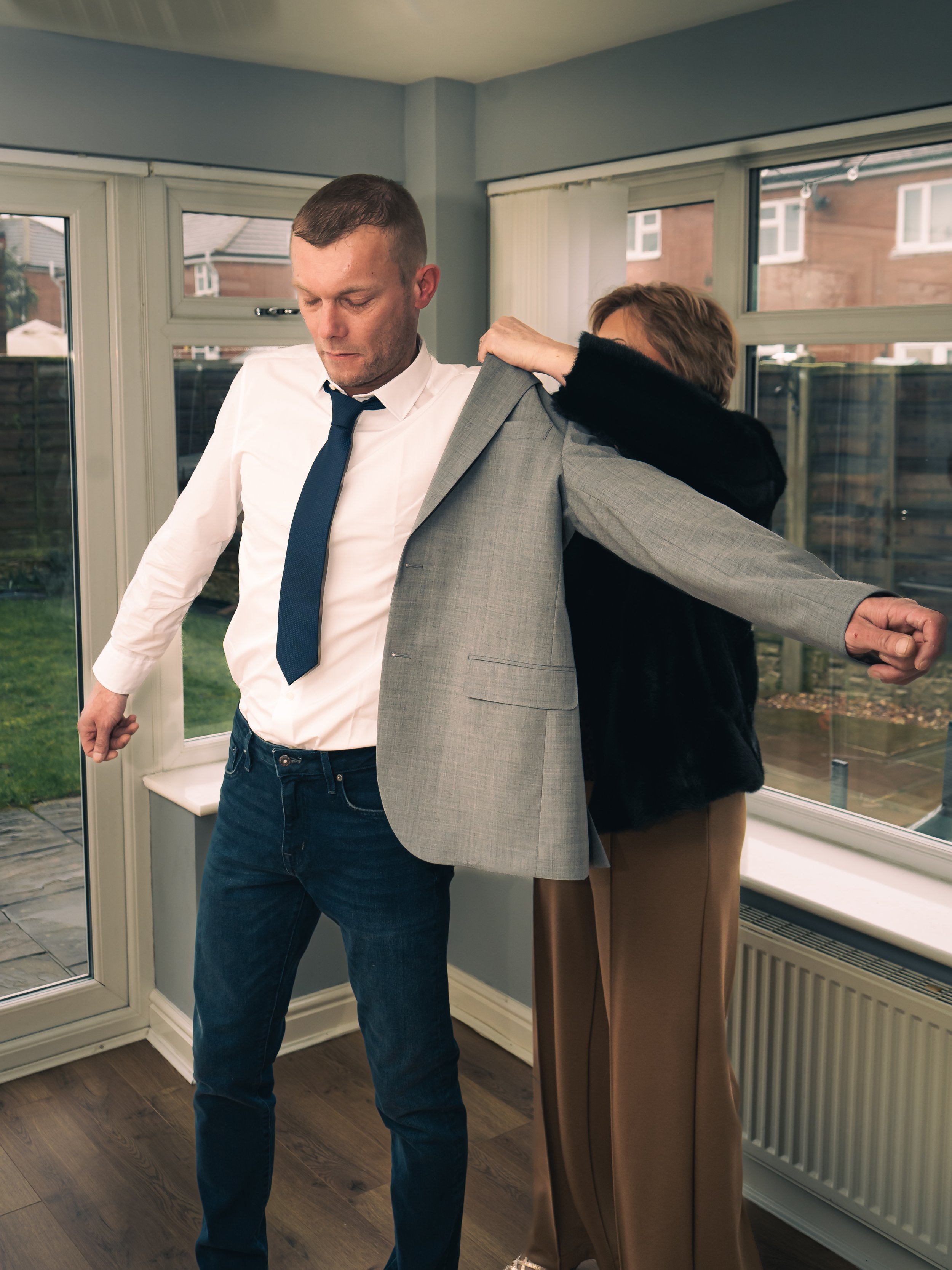 A man is trying on a light gray suit jacket with the help of a woman inside a bright room with large windows, a radiator, and a view of neighboring houses and a backyard.