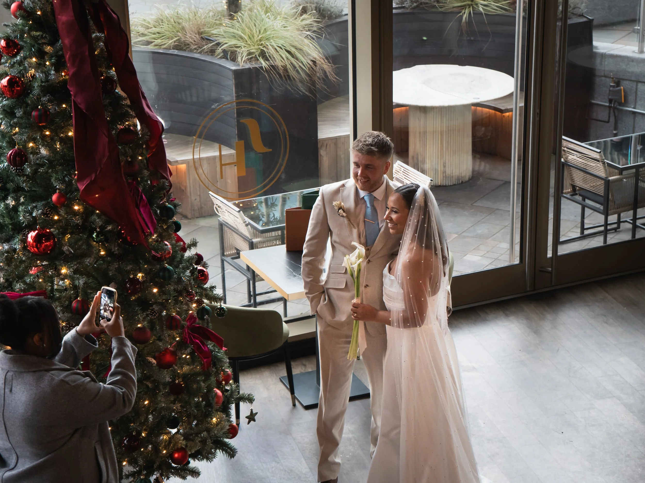 A bride and groom are standing near a decorated Christmas tree, taking a photo of the bride with a woman holding a phone, inside a modern building with large windows and outdoor seating.