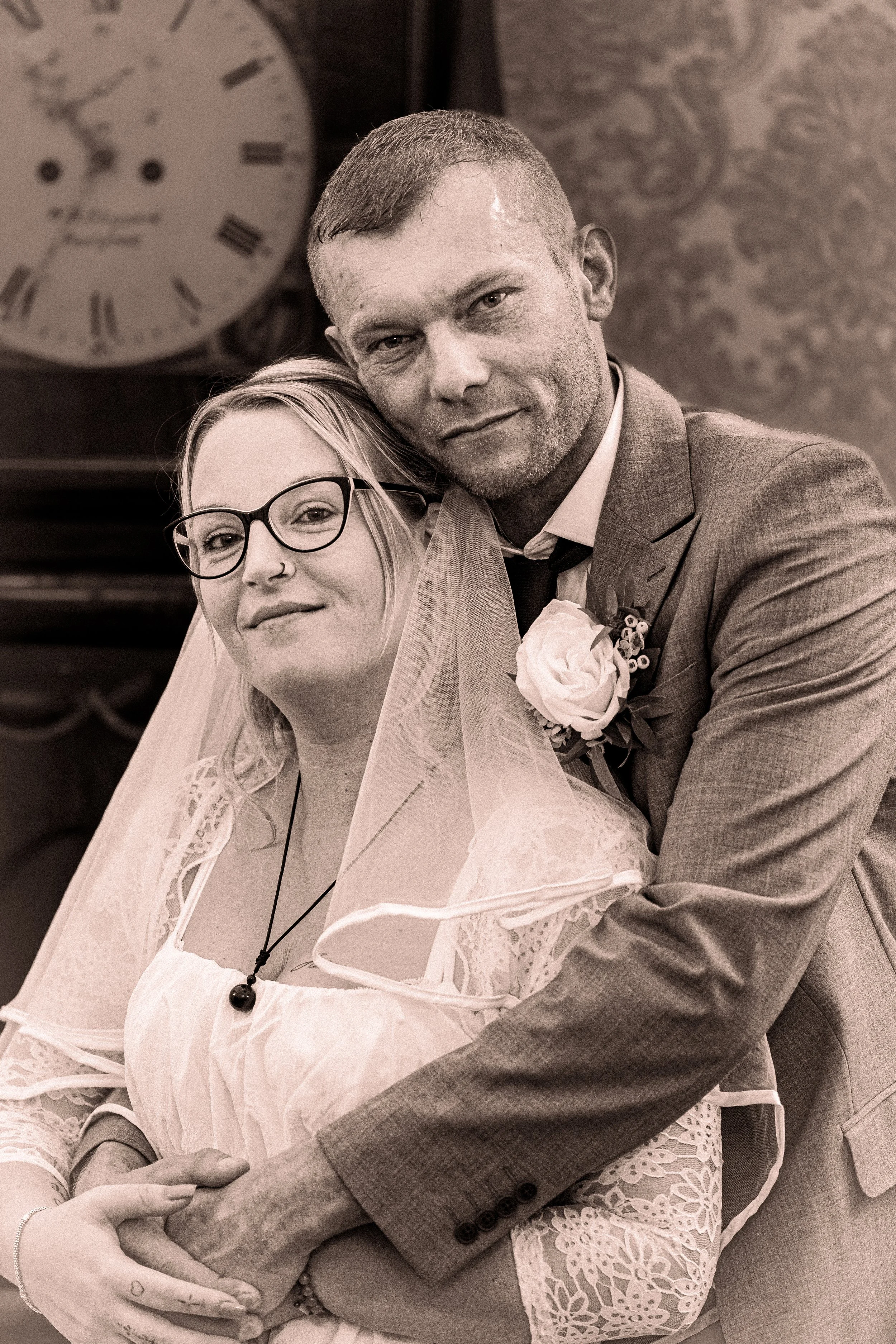 A black and white photograph of a couple, likely newlyweds, embracing closely. The woman has light hair, glasses, and a nose ring, and is wearing a lace dress and a veil. The man has short hair, a beard, and is wearing a suit with a boutonniere. They