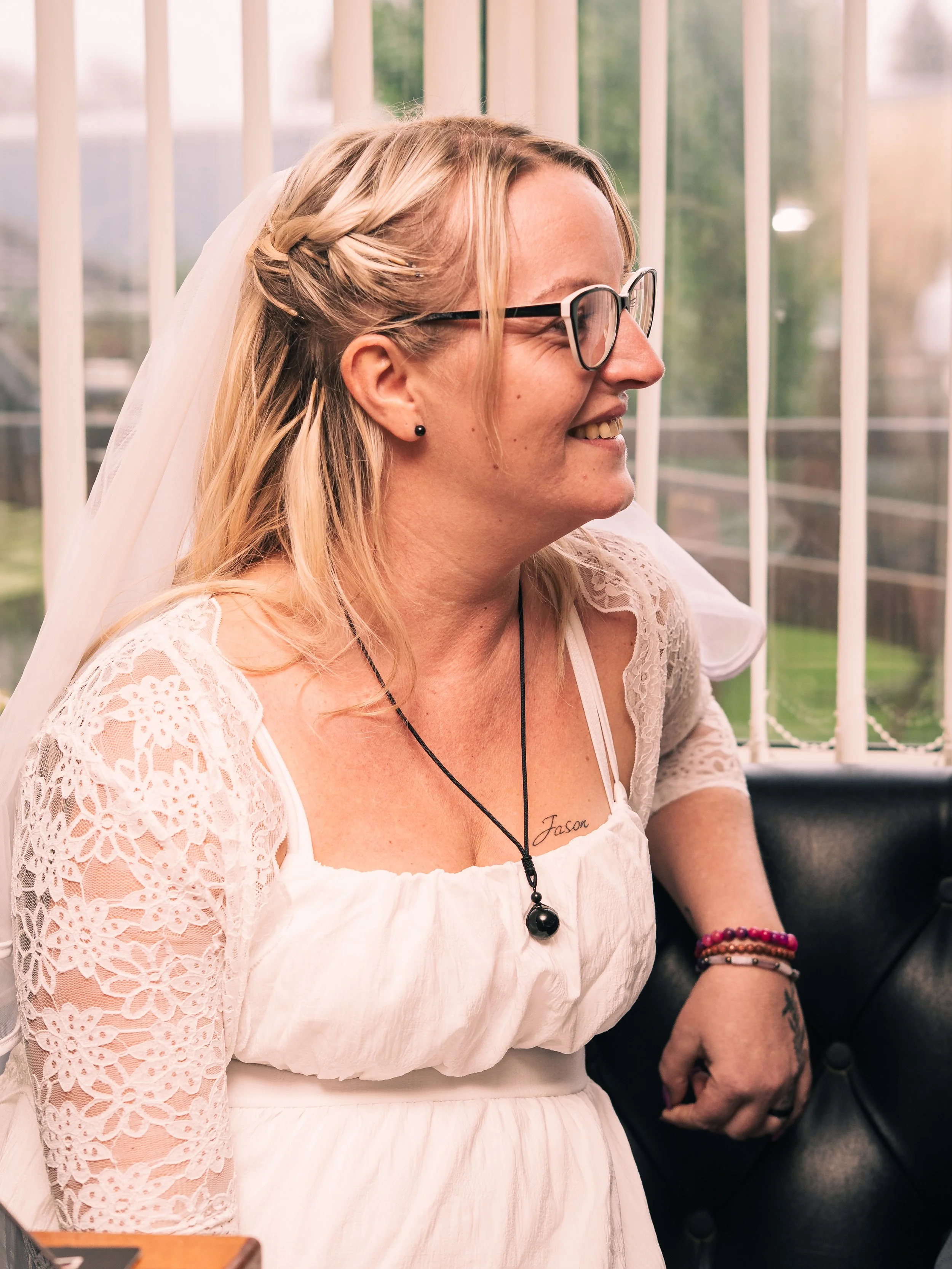 A woman wearing glasses and a white lace dress smiling while sitting on a black leather couch with a white veil and a tattoo reading 'Jason' visible on her chest.