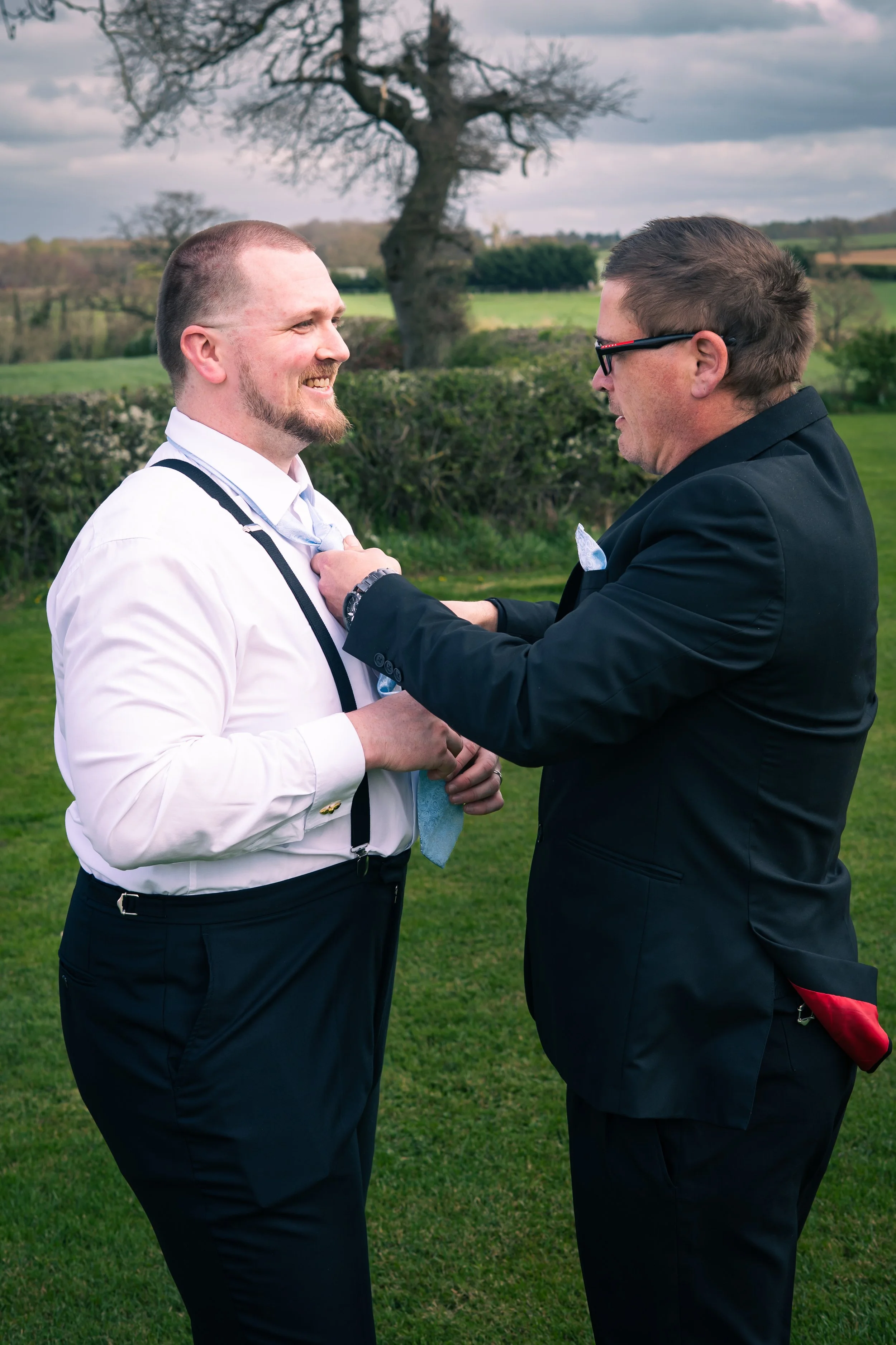 Two men in formal attire, one adjusting the other's tie outdoors on a grassy field, with trees and cloudy sky in the background.