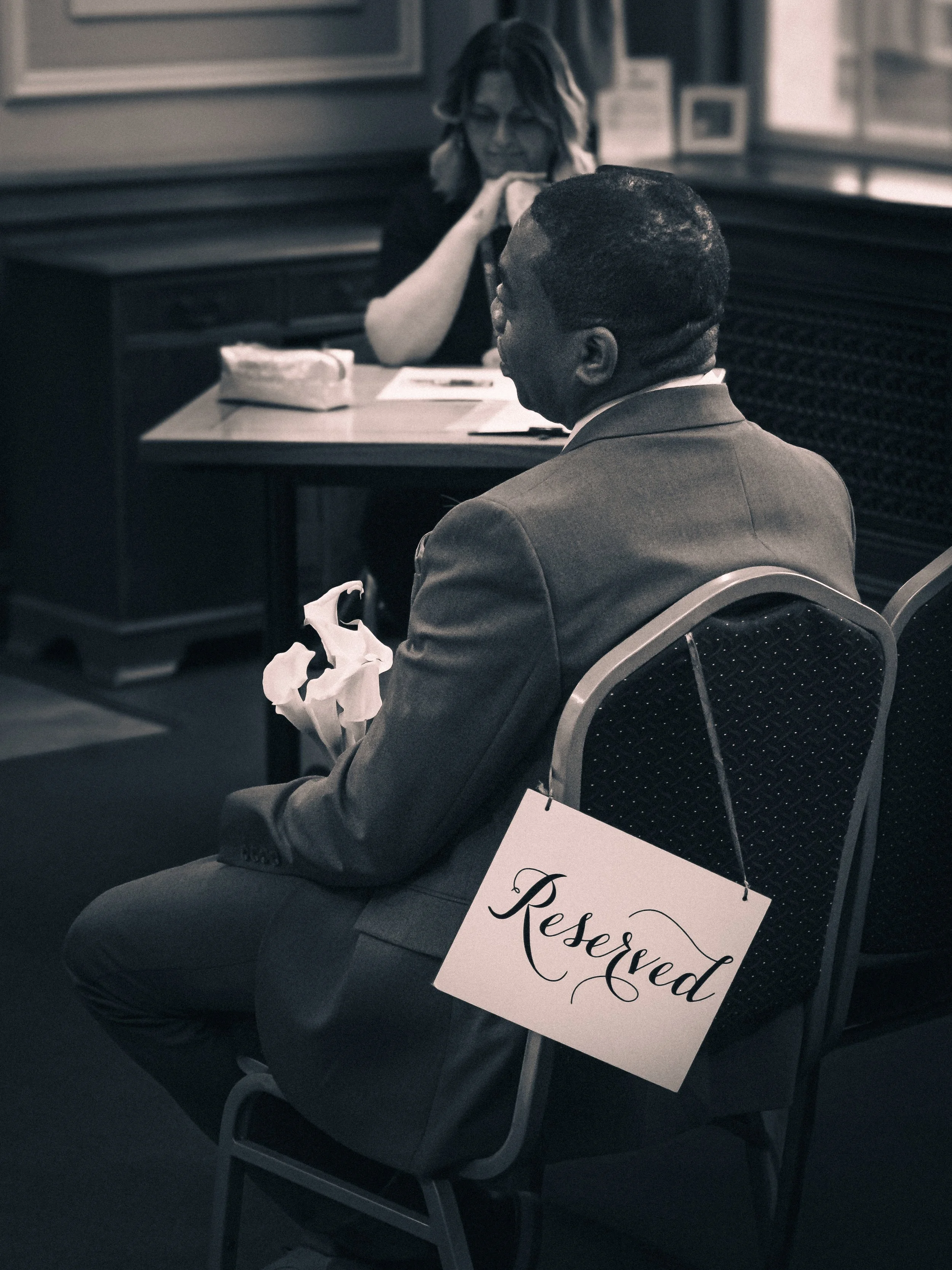 A man in a suit sitting in a chair with a 'Reserved' sign hanging from it, in a meeting room. A woman sits at a desk in front of him, smiling and observing.