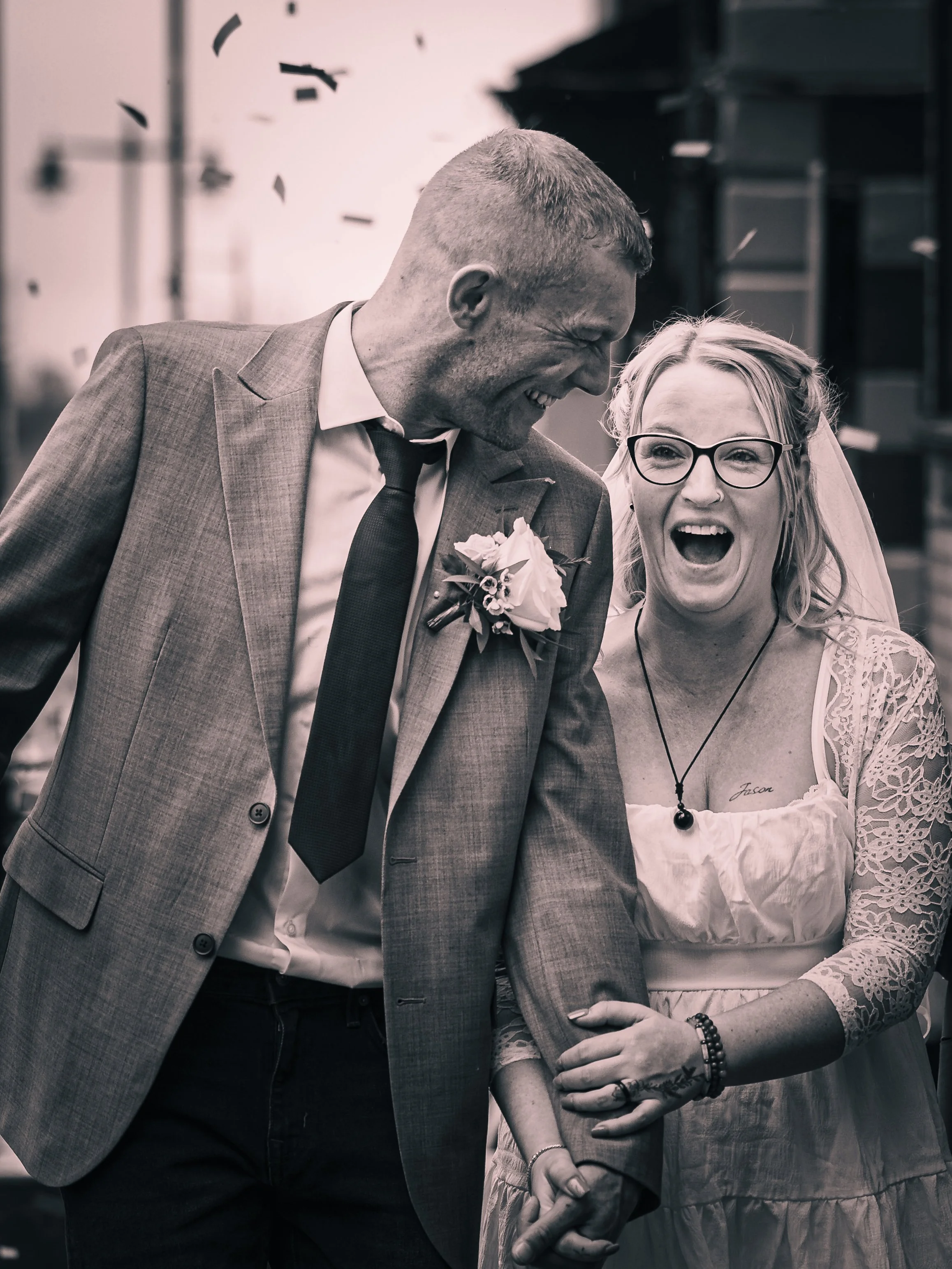 A black-and-white photo of a couple celebrating, with the man in a suit and the woman in a wedding dress, both smiling and holding hands, during a joyful moment at a wedding or celebration.