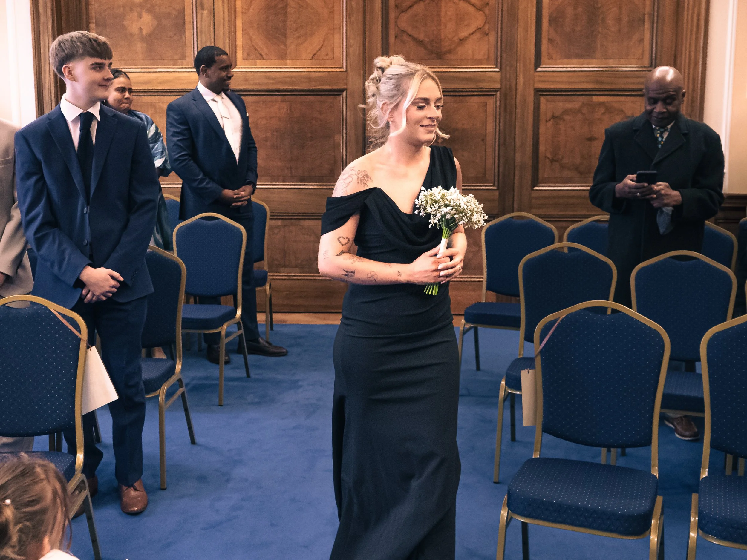A woman in a black dress holding a bouquet of flowers walking through a room with people dressed formally, some sitting and some standing. The room has wood-paneled walls and blue chairs with gold trim.