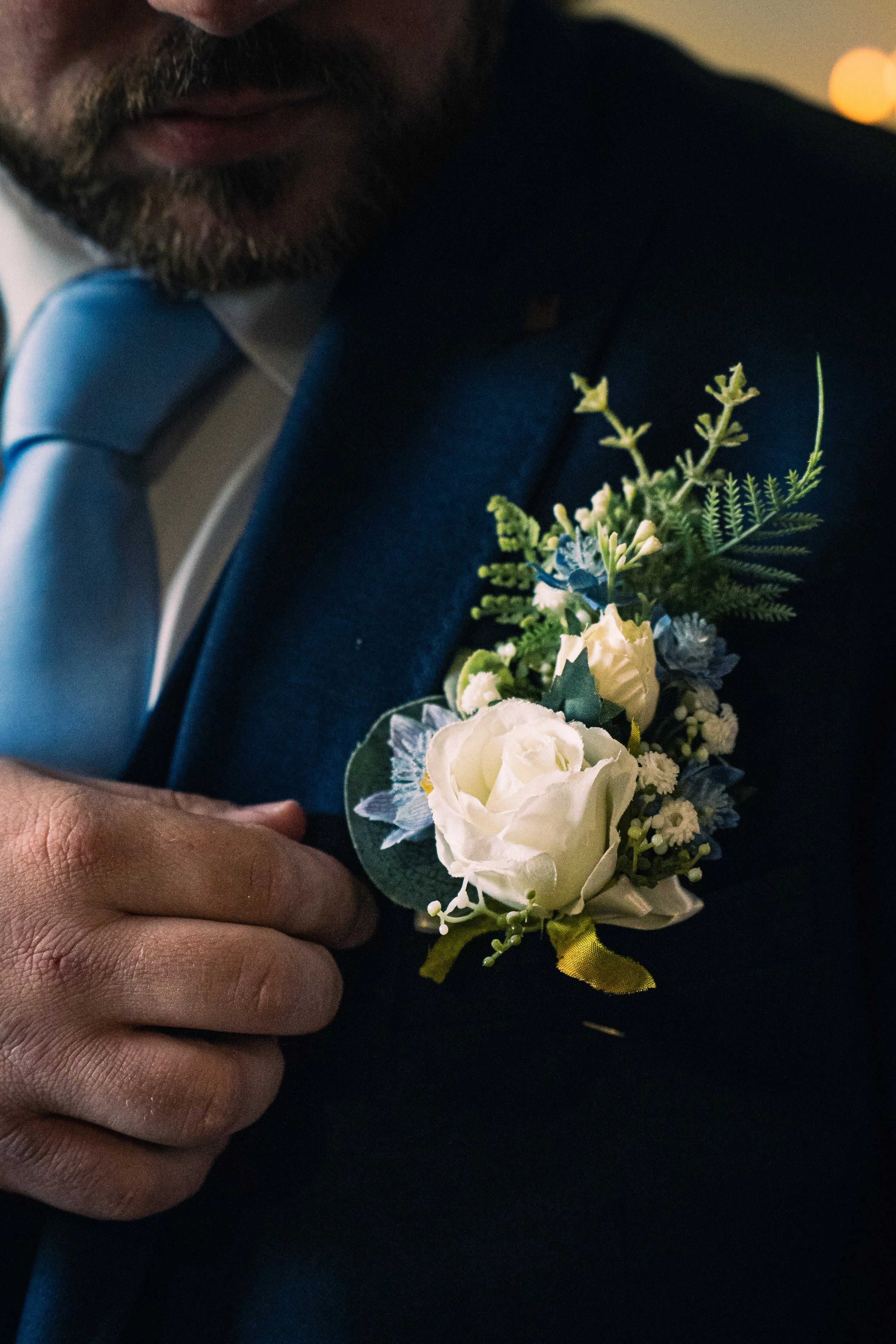 A man adjusts his suit jacket with a boutonniere pinned to the lapel, consisting of white roses, small white flowers, green leaves, and fern-like foliage, in a close-up shot.