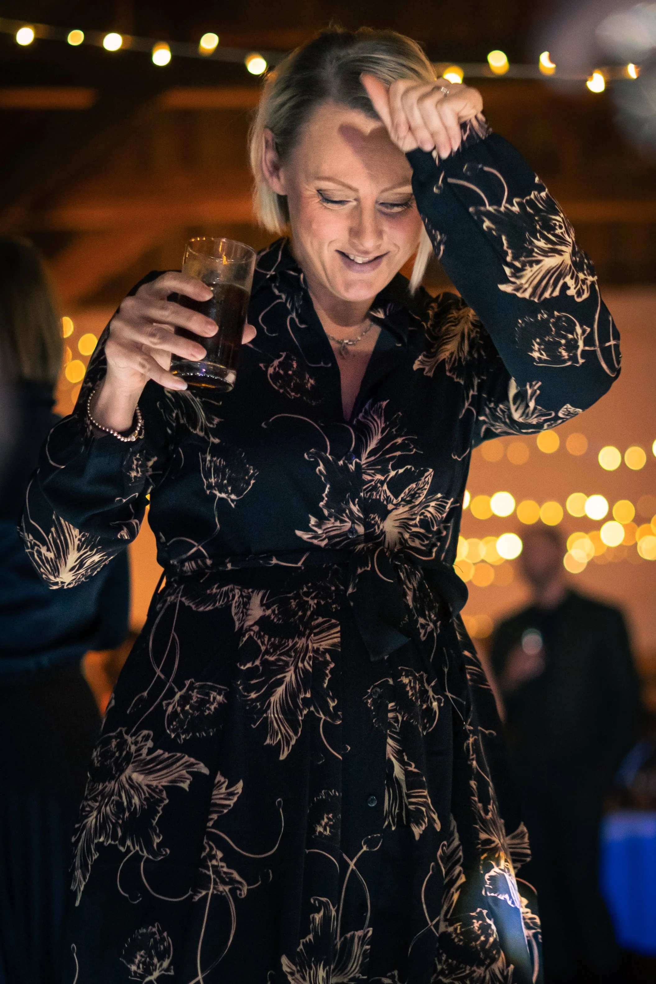 A woman smiling and dancing while holding a drink at a party, with blurred lights in the background.