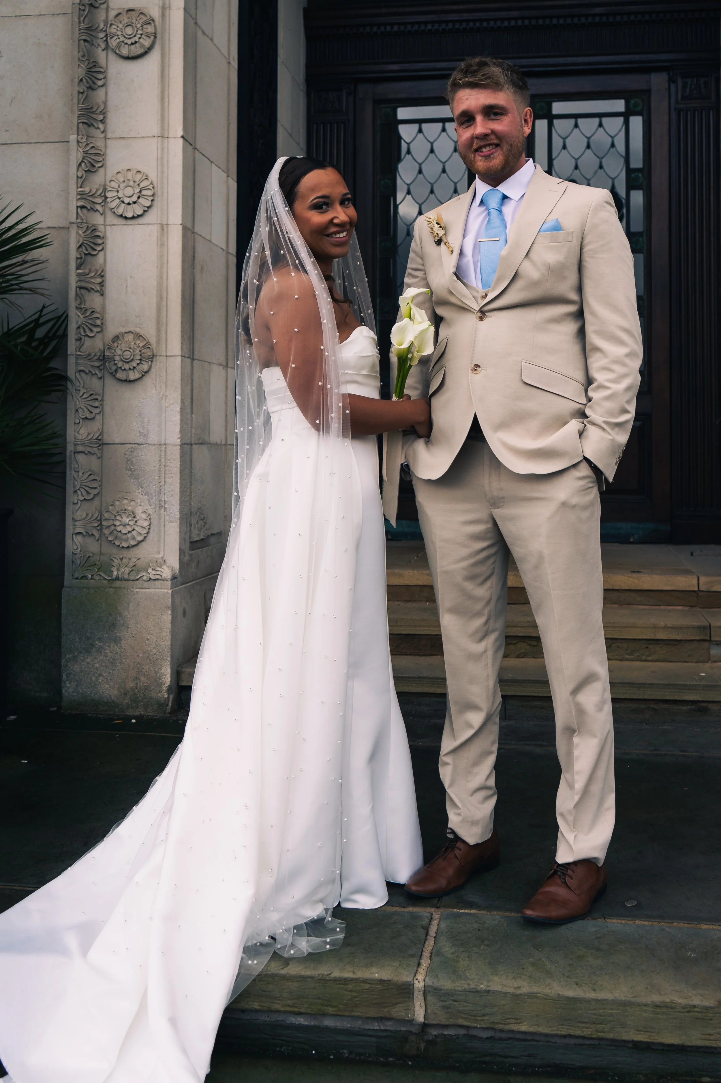 Bride and groom standing outside a building, smiling at the camera. The bride wears a white strapless wedding dress with a long train and a veil, holding a bouquet of white calla lilies. The groom wears a light beige suit with a white shirt and a lig