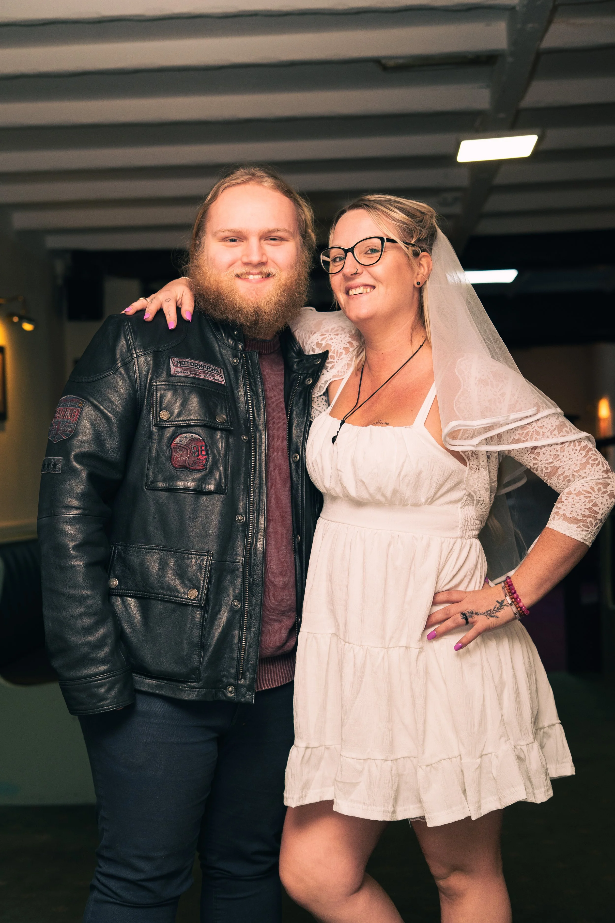 A happy man with long blond hair and a beard wearing a black leather jacket standing next to a smiling woman with blond hair, glasses, and a white dress, wearing a veil, in an indoor setting.