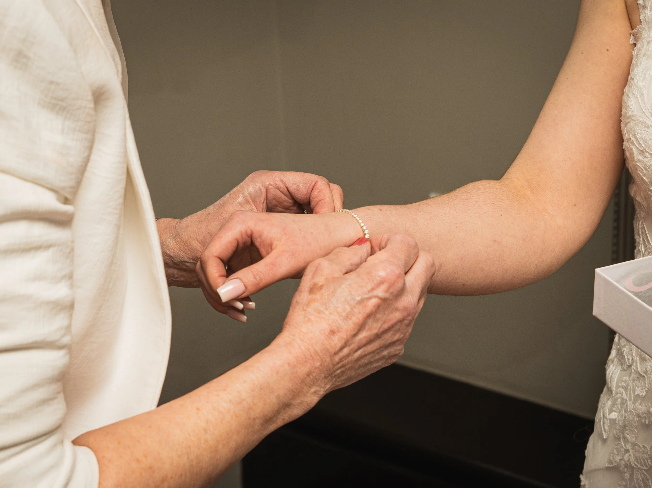 Close-up of an elderly woman placing a wedding ring on a young woman's finger. The young woman is wearing a lace dress, and the elderly woman is wearing a cream-colored outfit.