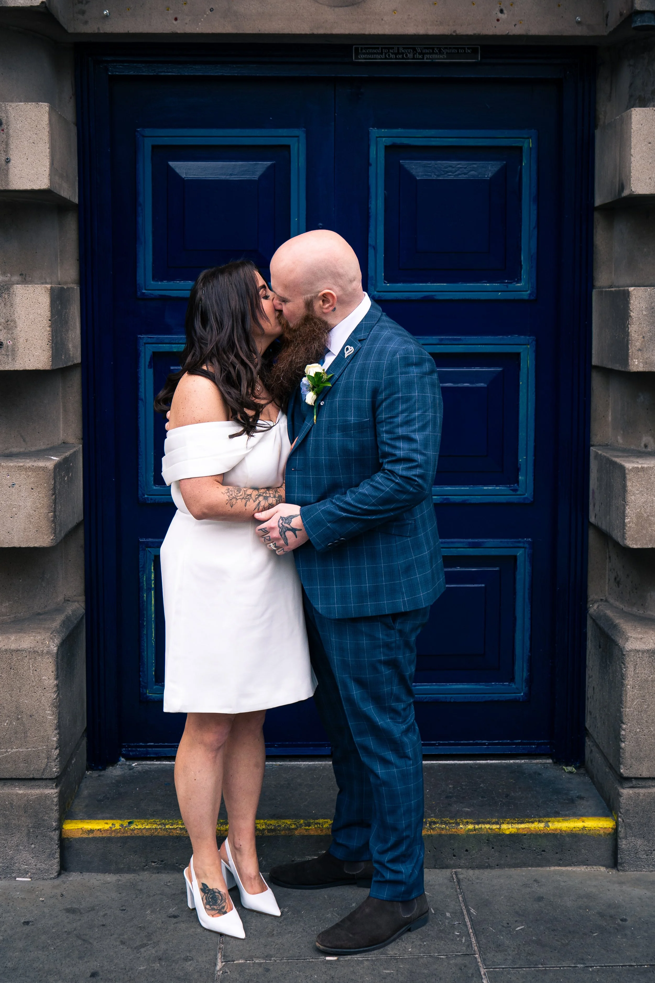 A bride and groom sharing a kiss in front of a blue door. The bride wears a white off-shoulder dress and white heels with tattoos on her arms and foot. The groom wears a blue plaid suit with black shoes.