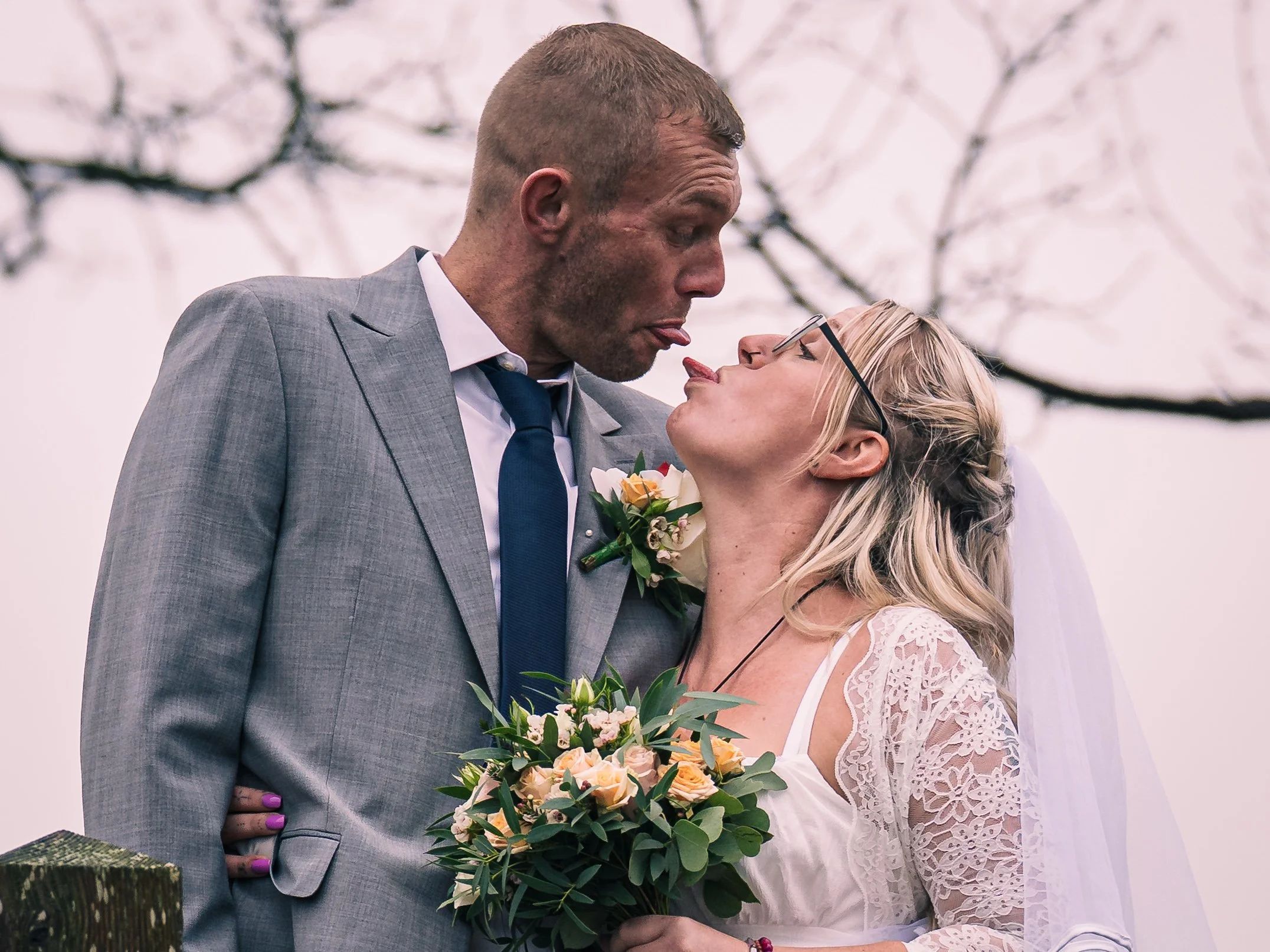 A bride and groom sharing a close moment outdoors during their wedding, with the groom wearing a gray suit and the bride in a white lace dress holding a bouquet of flowers.