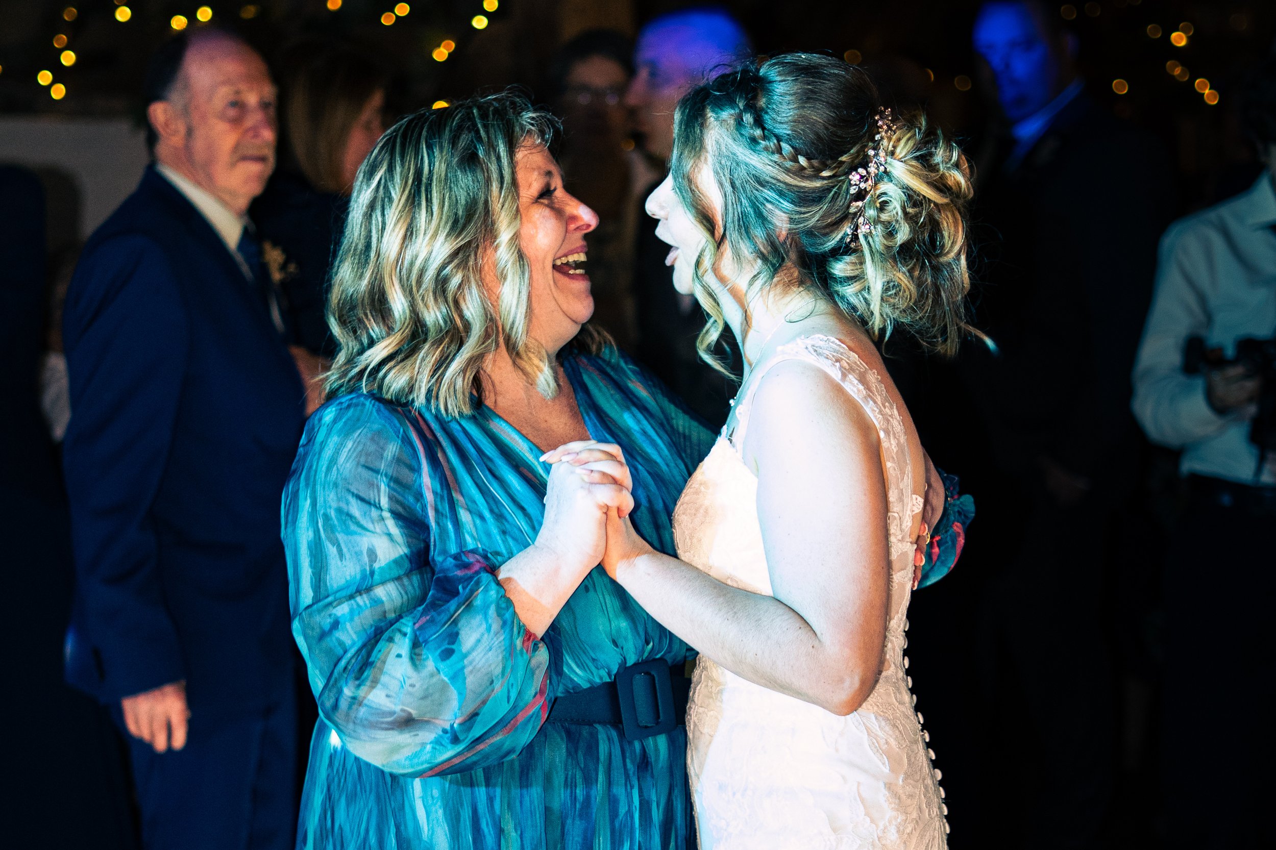 A woman and a bride dancing happily at a wedding reception. The woman is wearing a blue dress with long sleeves, and the bride has a wedding gown with lace details. Other guests are visible in the background, some watching and some holding phones.