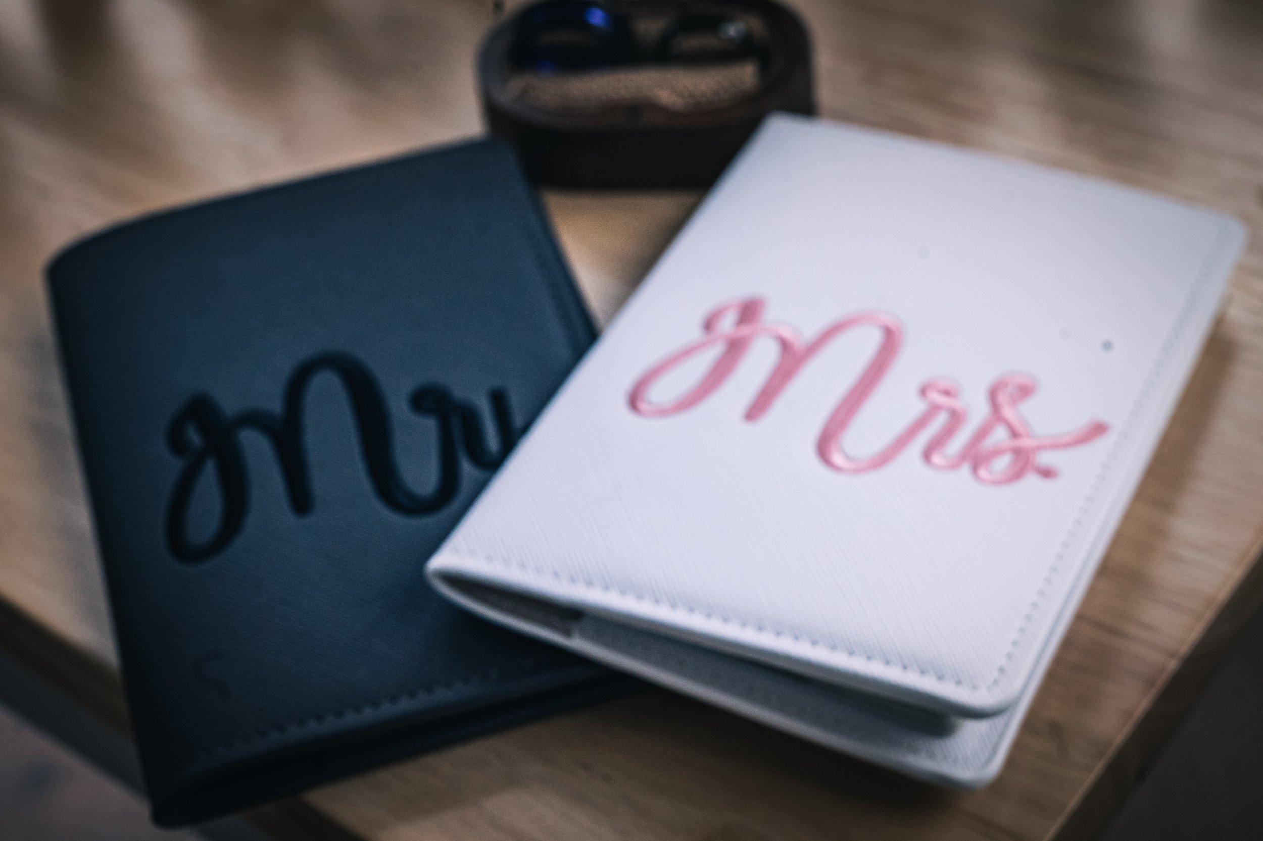 Two wallets, one black and one white, with the words 'Mrs' and 'Mrs' embroidered on them, and a black wristwatch on a wooden surface.