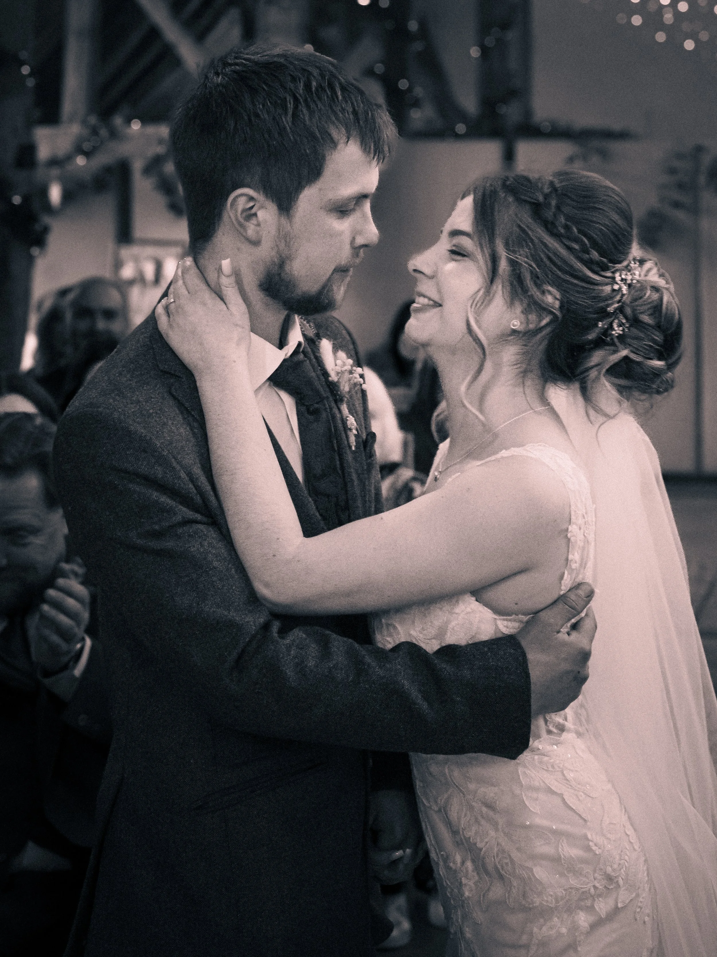 A bride and groom share a dance at their wedding reception, looking into each other's eyes with smiles, surrounded by guests.