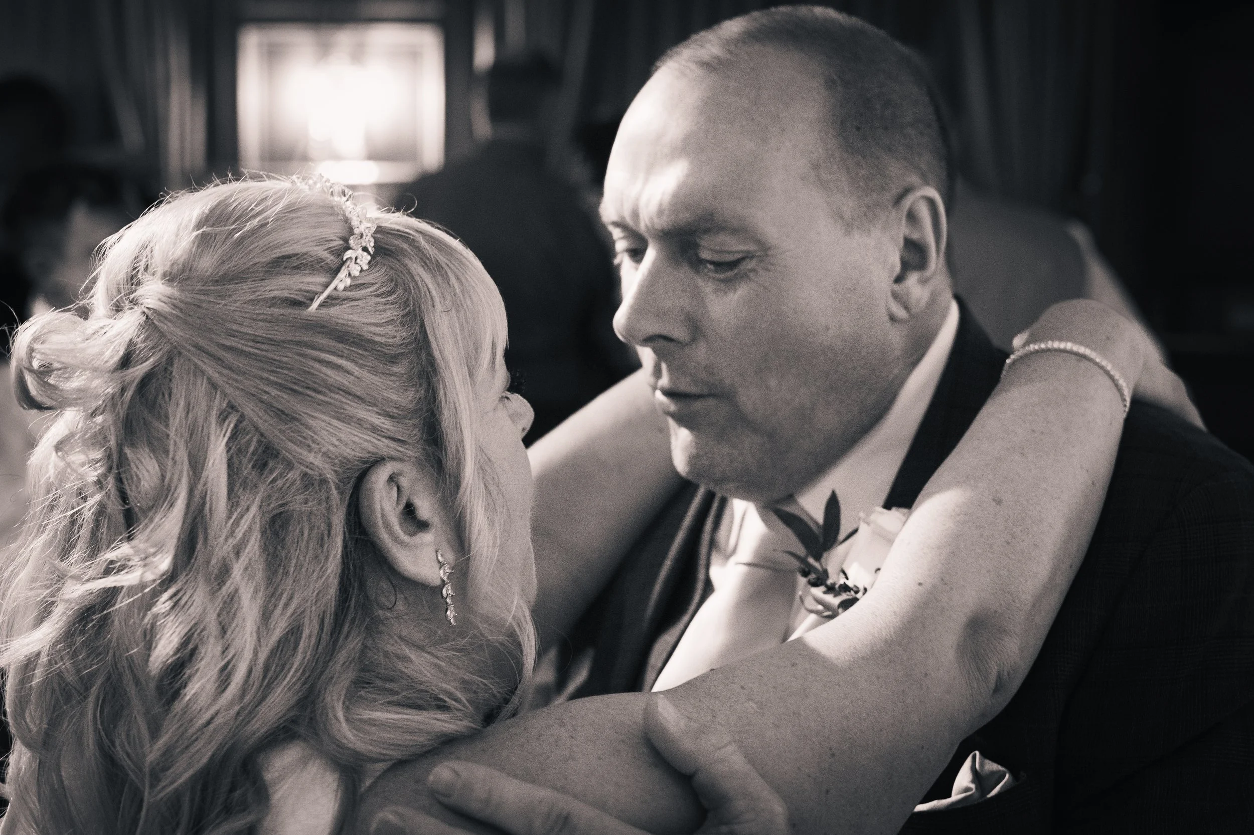A black and white photo of a couple at a wedding reception, looking into each other's eyes. The woman has blonde wavy hair and jewelry, and the man is dressed in a suit with a boutonniere.