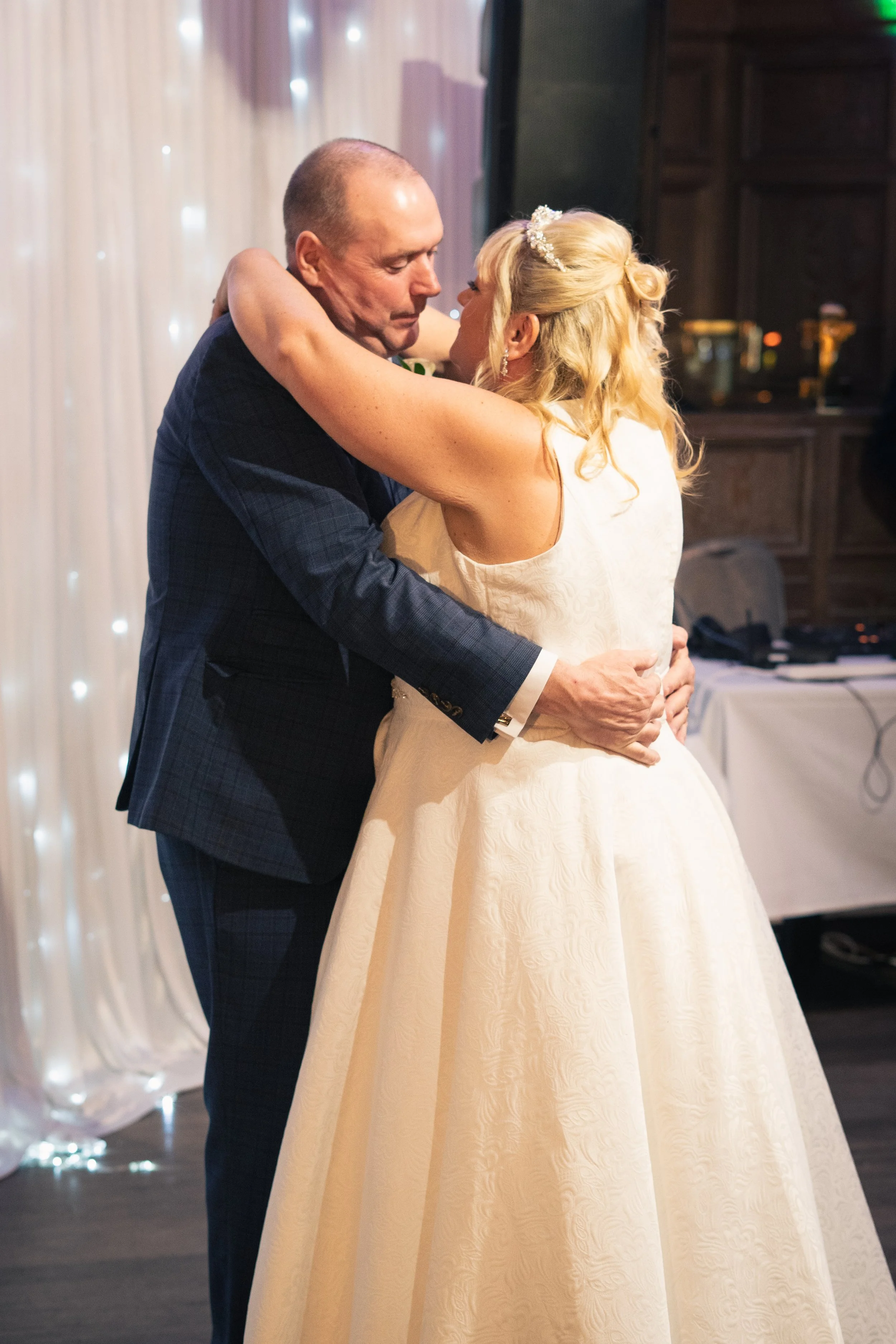 A bride and an older man, possibly her father, sharing a dance at a wedding reception. The bride is in a white gown with a floral pattern and a headband, and the man is in a dark suit with a checked pattern. They are embracing each other closely, wit